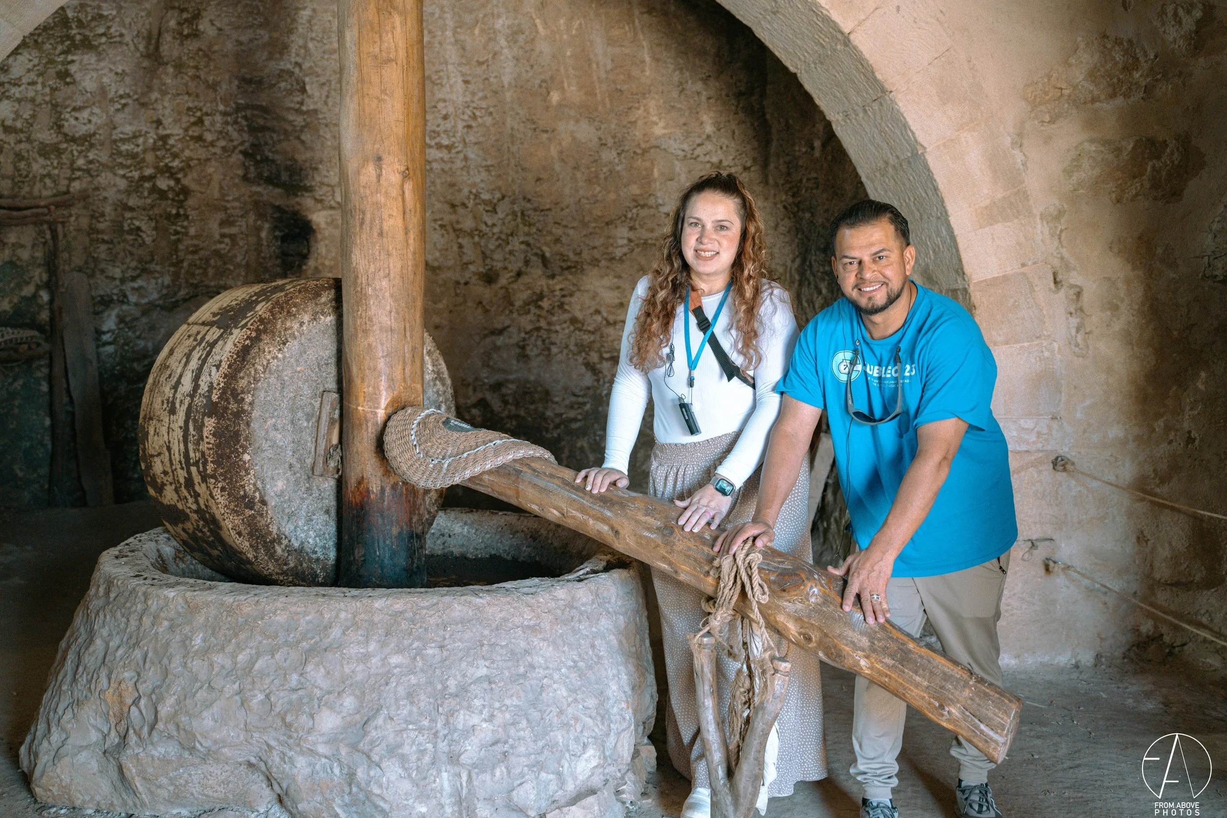 Two people, a woman and a man, standing inside an old stone mill with a giant stone wheel, holding a wooden and rope handle.