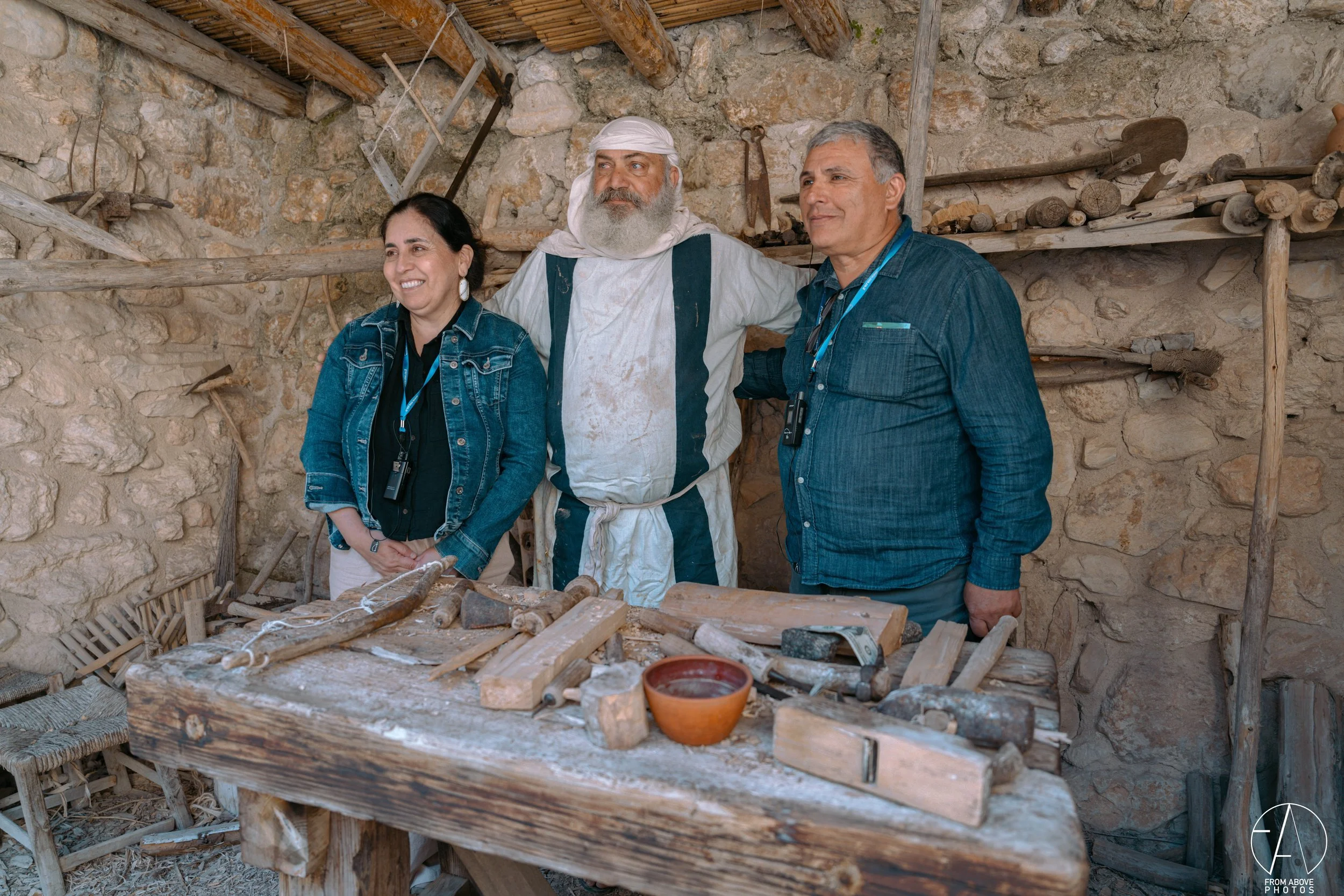 Three people standing inside a rustic woodworking workshop. One man is dressed in historical clothing, while two others are in modern casual attire with cameras hung around their necks. They are smiling and leaning on a wooden workbench filled with w