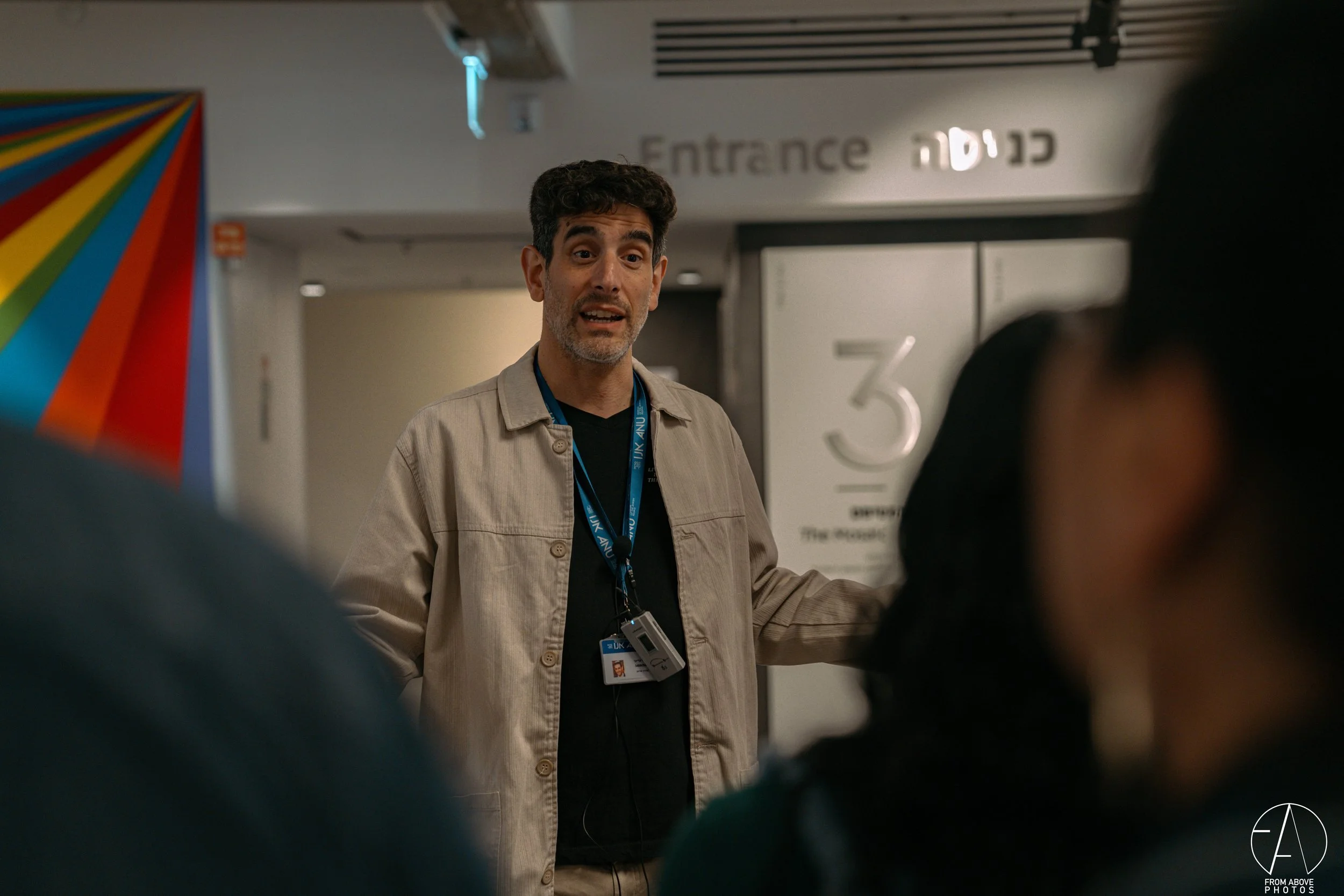 A man with dark hair and a beard, wearing a beige jacket and a lanyard, standing in front of an entrance sign at an airport.