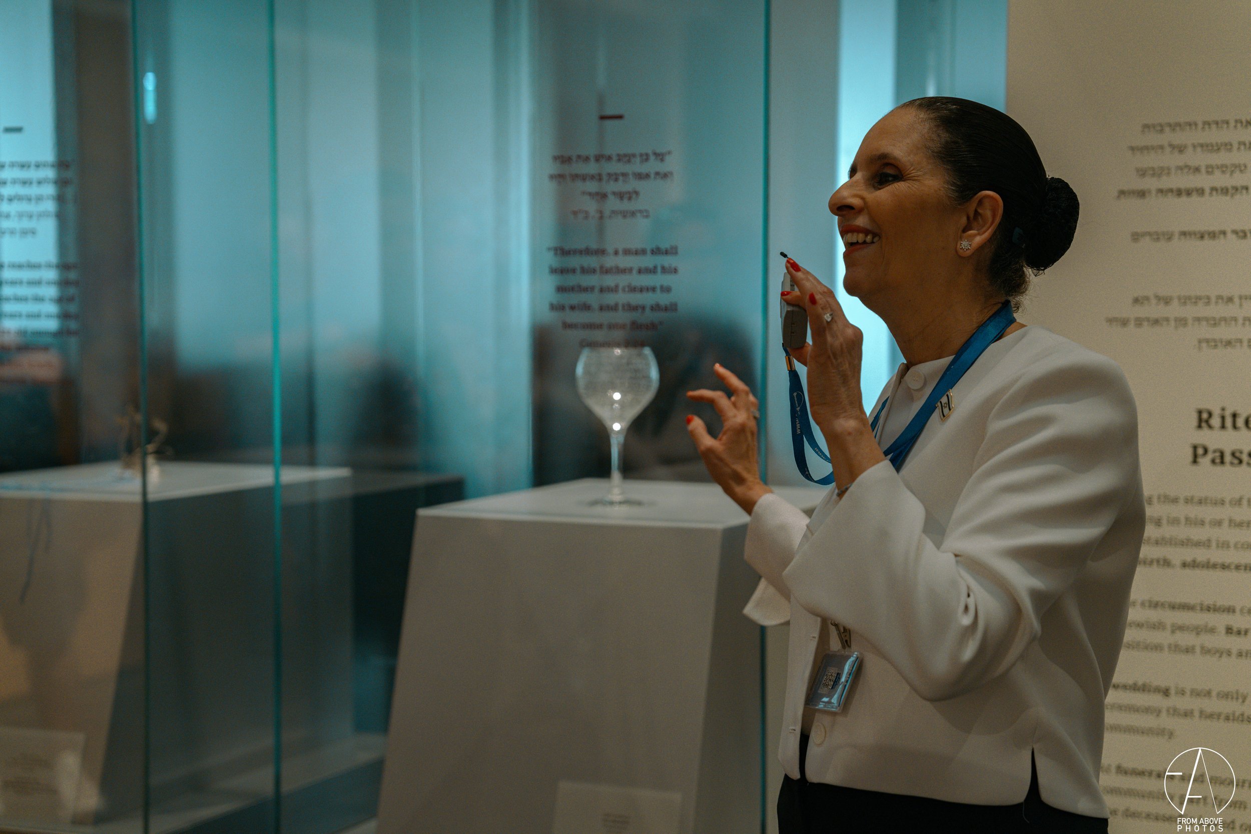 A woman with dark hair tied back, wearing a white blazer and a blue lanyard, is speaking into a microphone in a museum exhibit. Behind her are glass display cases with artifacts and informational text on the walls.