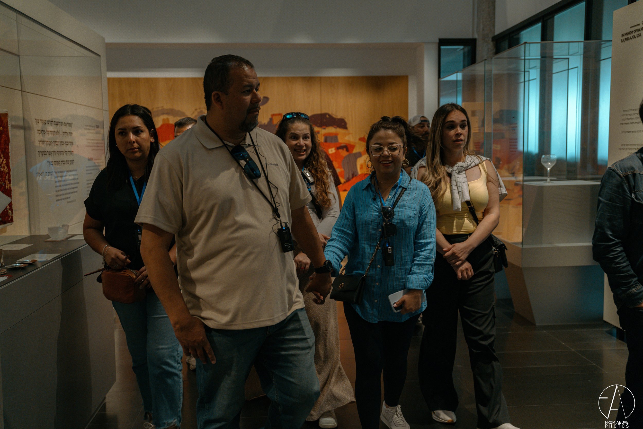 A group of people touring a museum exhibit, including men and women of diverse backgrounds, some wearing badges and holding small objects or phones, with display cases and informational panels in the background.