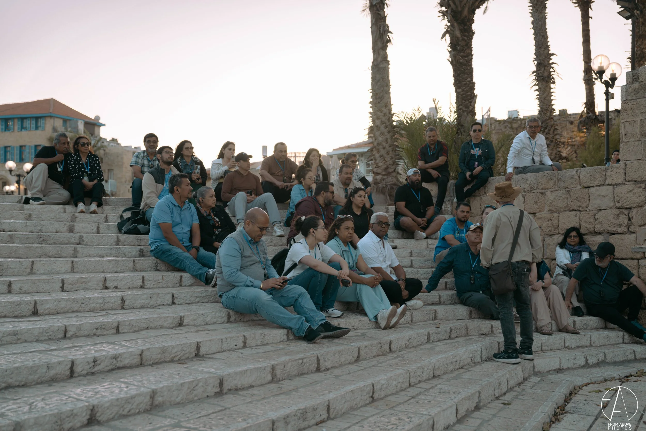 A group of people sitting on stone steps outdoors, listening to a man with a hat and a beige jacket who is standing and speaking to them. There are tall palm trees and old stone walls in the background, suggesting a historic or scenic location during