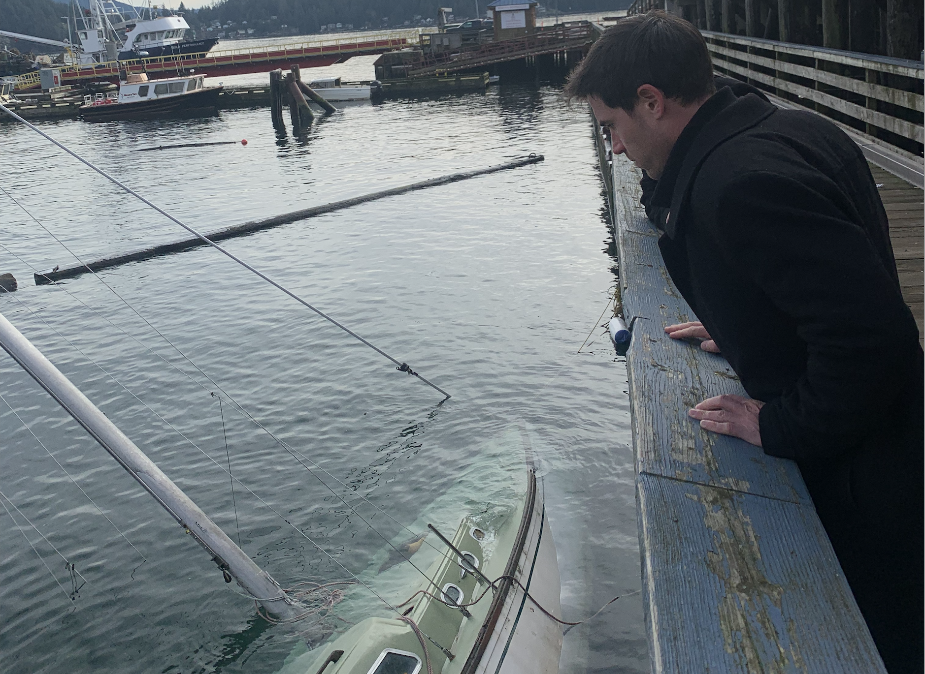 A man leaning over a dock looking into the water where a sailboat has capsized, with boats and a marina in the background.