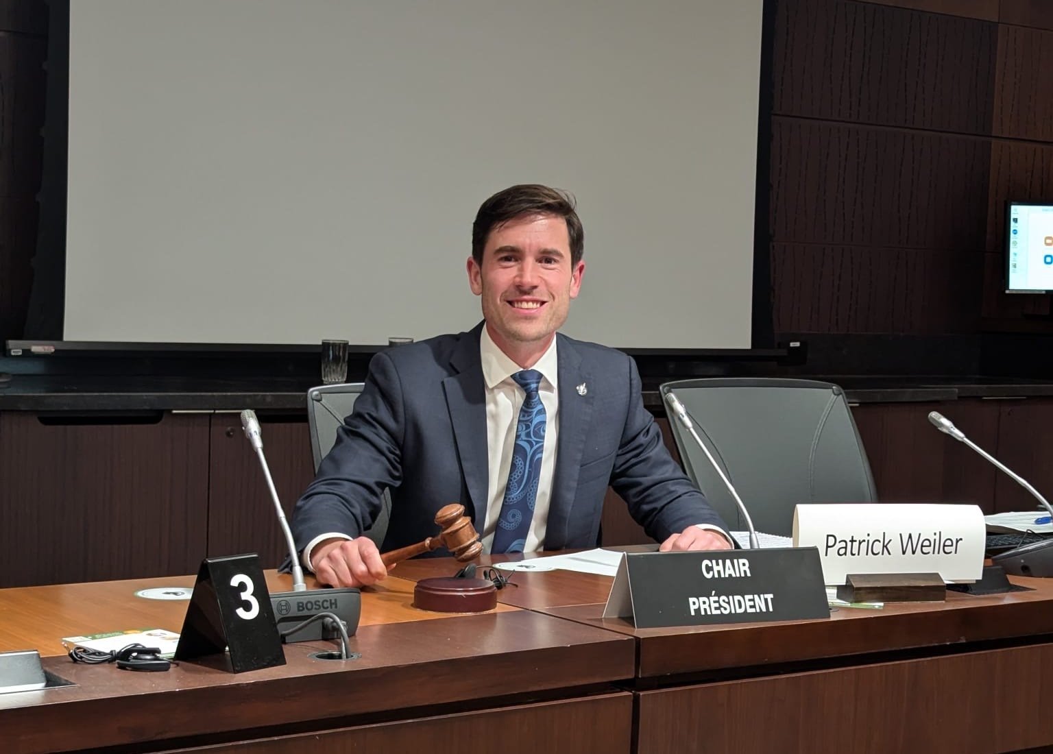 A man in a suit sitting at a conference table with a gavel in his hand. There are nameplates in front of him, one reads 'CHAIR PRÉSIDENT' and another 'Patrick Weiler.' The man is smiling, and there are microphones on the table. The background includes a blank screen and wood paneling.
