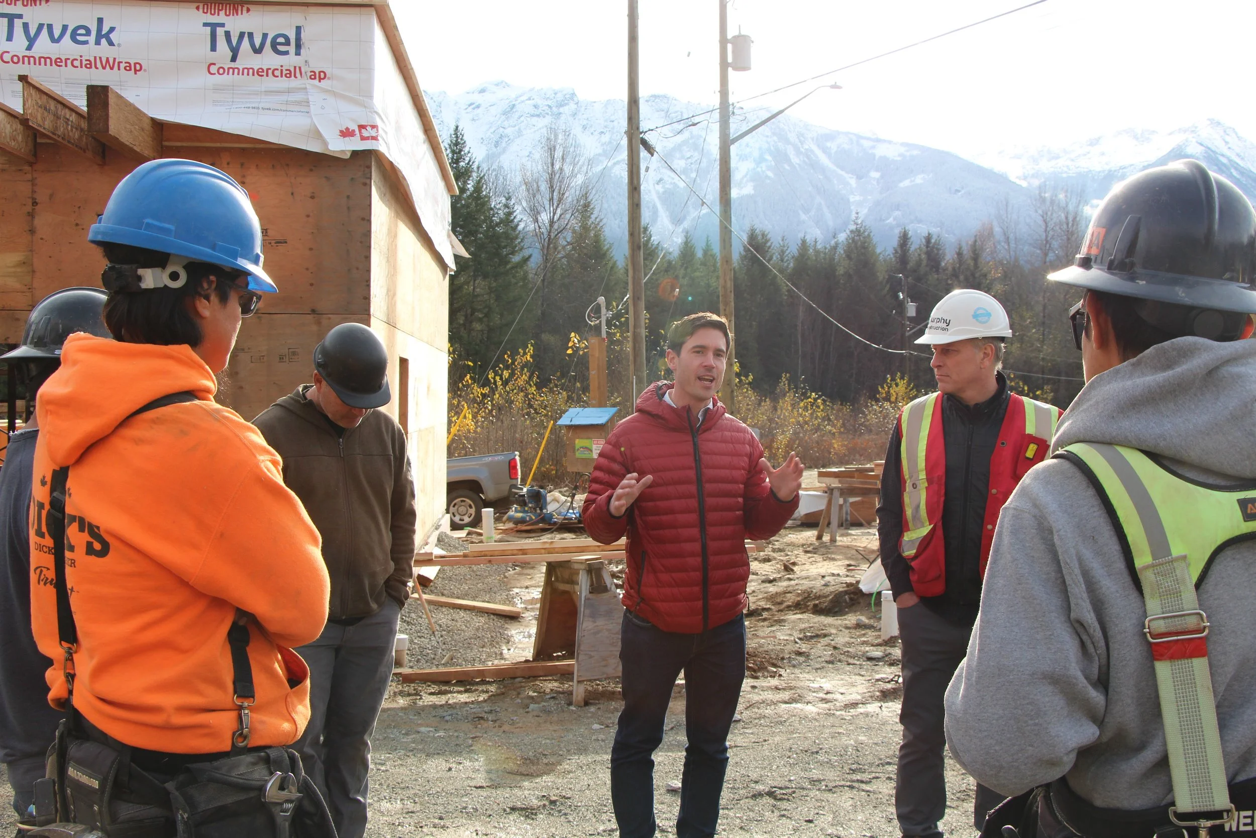 Group of construction workers and a man giving a talk at a building site with a wooden frame, electrical poles, and mountains in the background.
