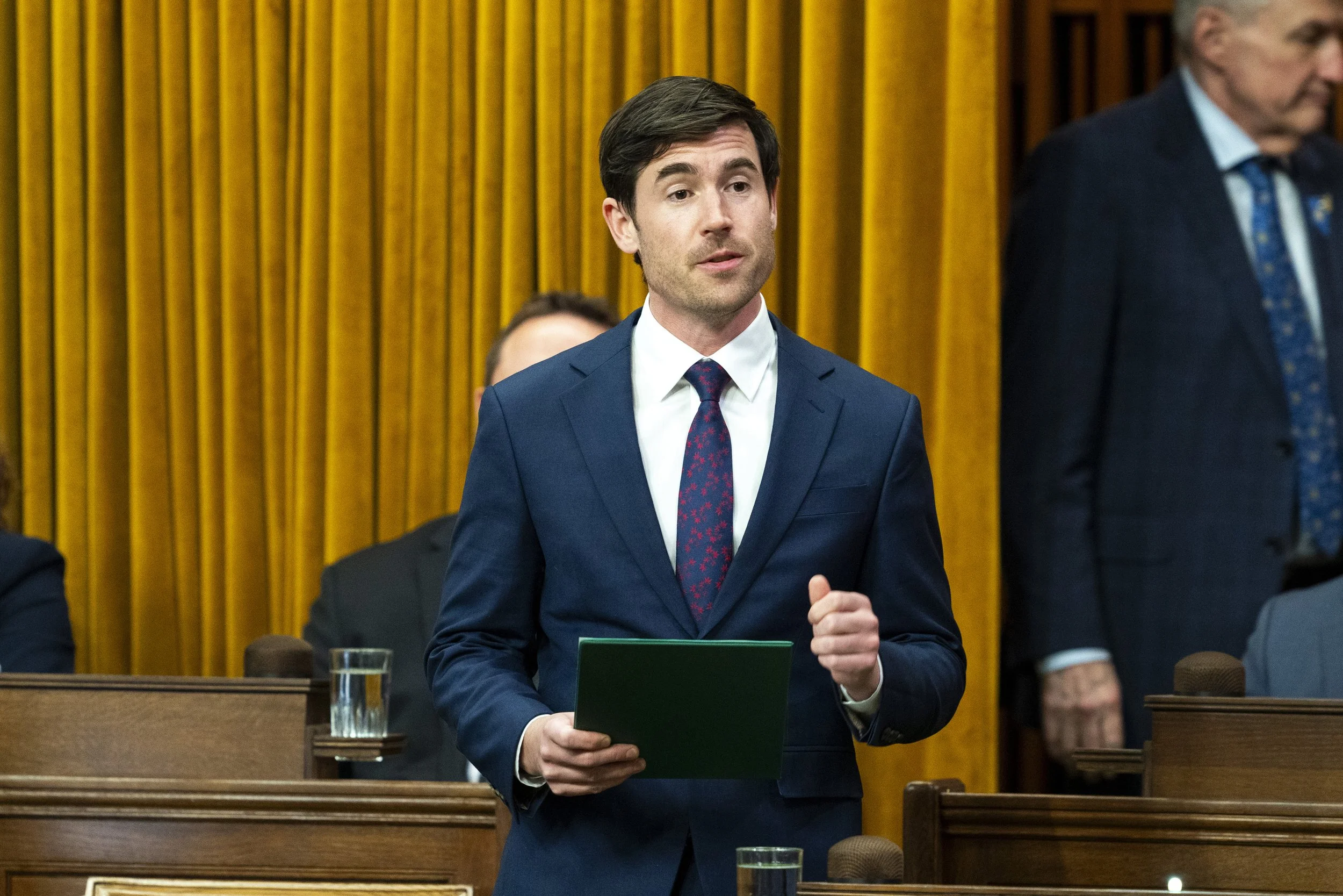 A man with dark hair in a navy blue suit, white shirt, and patterned red tie standing in a formal setting with wooden paneling and other people, some wearing suits, behind him. He is holding a green folder and a glass of water is on the desk in front of him.