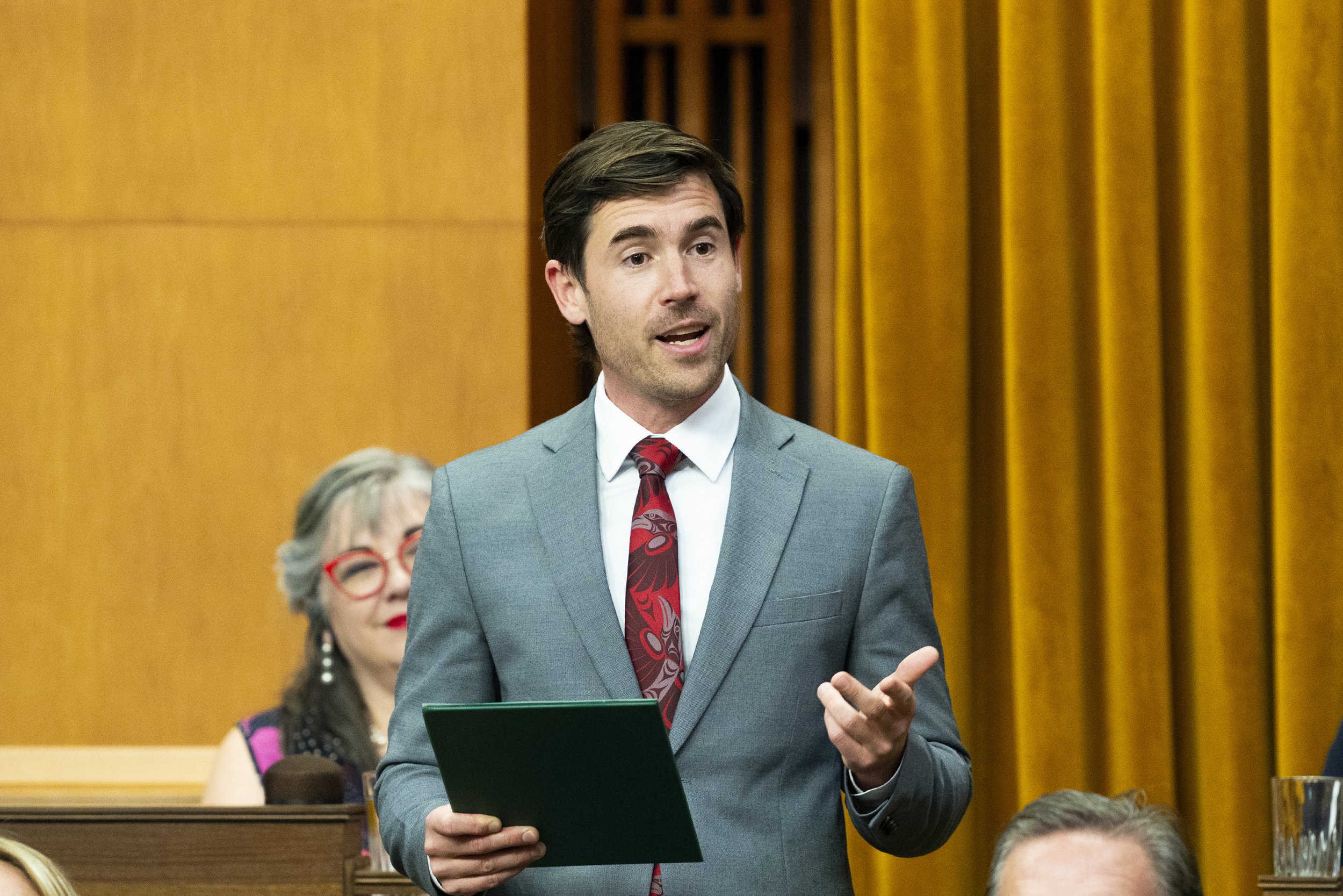 A man with dark hair wearing a gray suit, white shirt, and red patterned tie speaking in a formal setting, holding a green folder, with a woman with curly hair and red glasses sitting behind him, and a wood-paneled wall in the background.