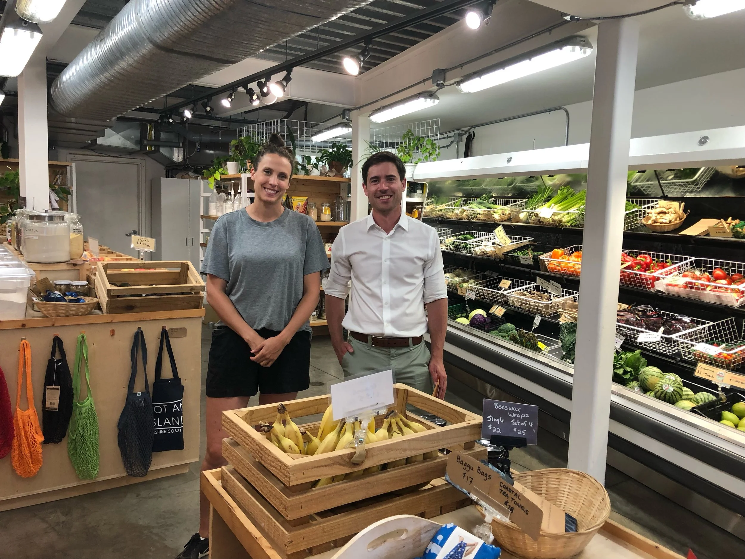 Two people, a woman in a casual grey t-shirt and a man in a white dress shirt, are standing inside a grocery store near produce and grocery items. The store has an industrial ceiling with exposed ductwork and is well-lit with fluorescent lighting. There are various fresh vegetables and bananas visible behind them.