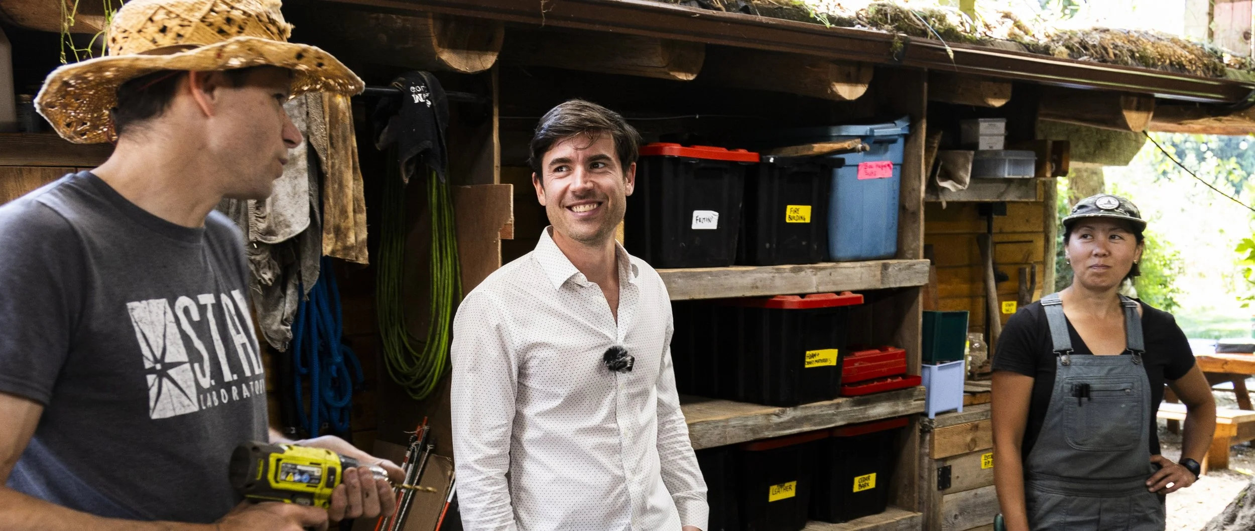 Three people standing outdoors in front of a wooden shed with storage bins and hanging tools, engaged in conversation. One man is wearing a straw hat and a graphic t-shirt, holding a small electronic device, another man in a white shirt is smiling, and a woman wearing a cap and overalls is looking on.