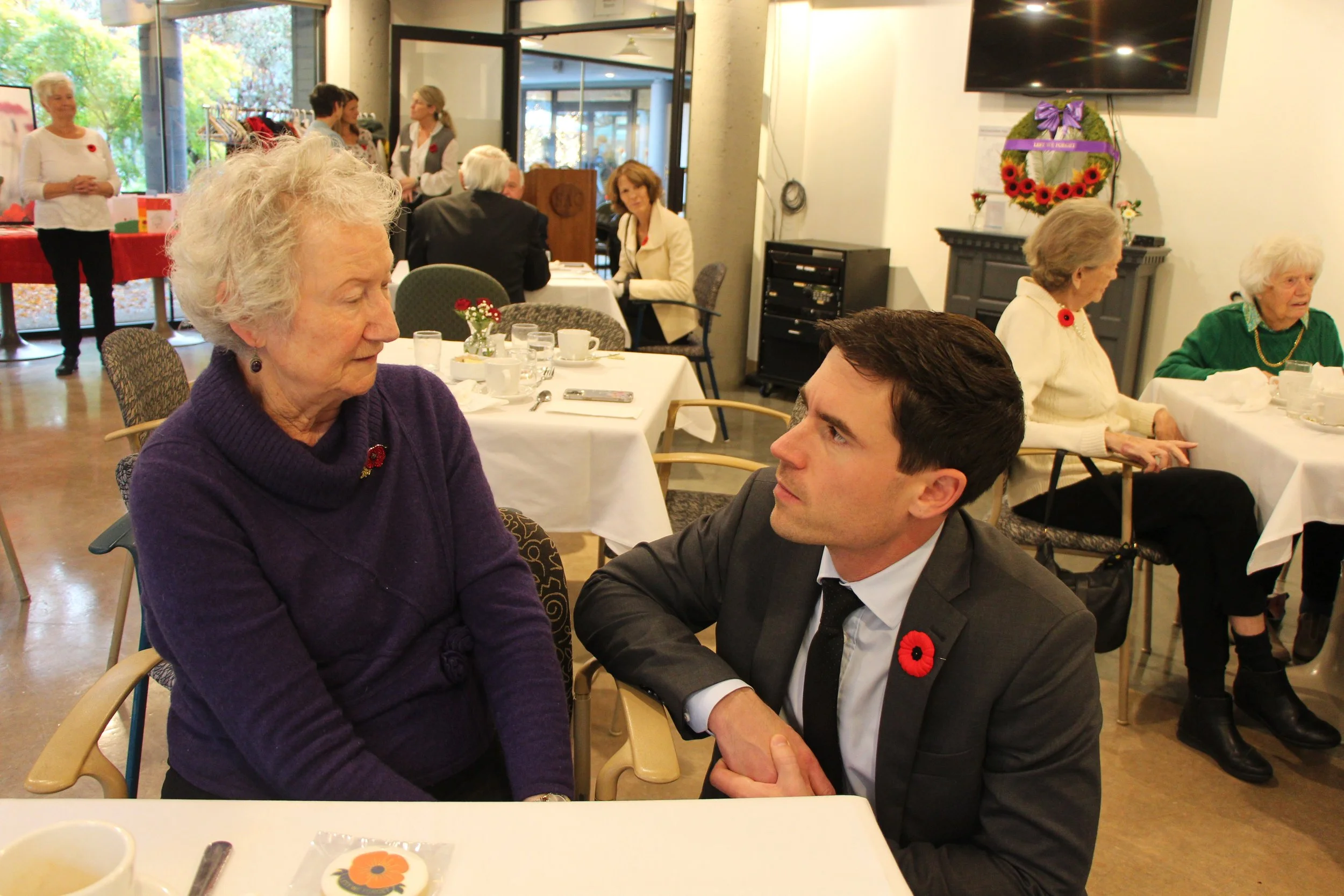 An elderly woman and a young man are sitting at a dining table, holding hands and looking at each other fondly, in a room filled with other people, some sitting and some standing, all wearing poppies for Remembrance Day. The elderly woman is wearing a purple sweater and the young man is dressed in a suit with a poppy pin on his lapel.