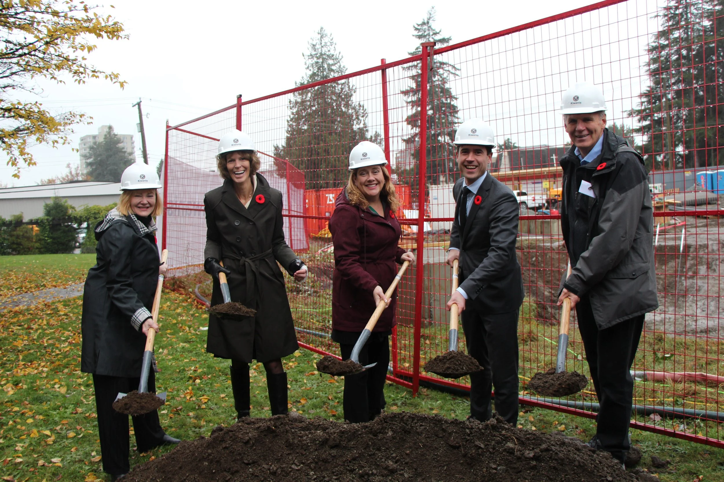 Five people in formal attire and hard hats holding shovels with dirt, standing in front of a red construction fence, at a ground-breaking ceremony in a park with trees and fallen leaves.