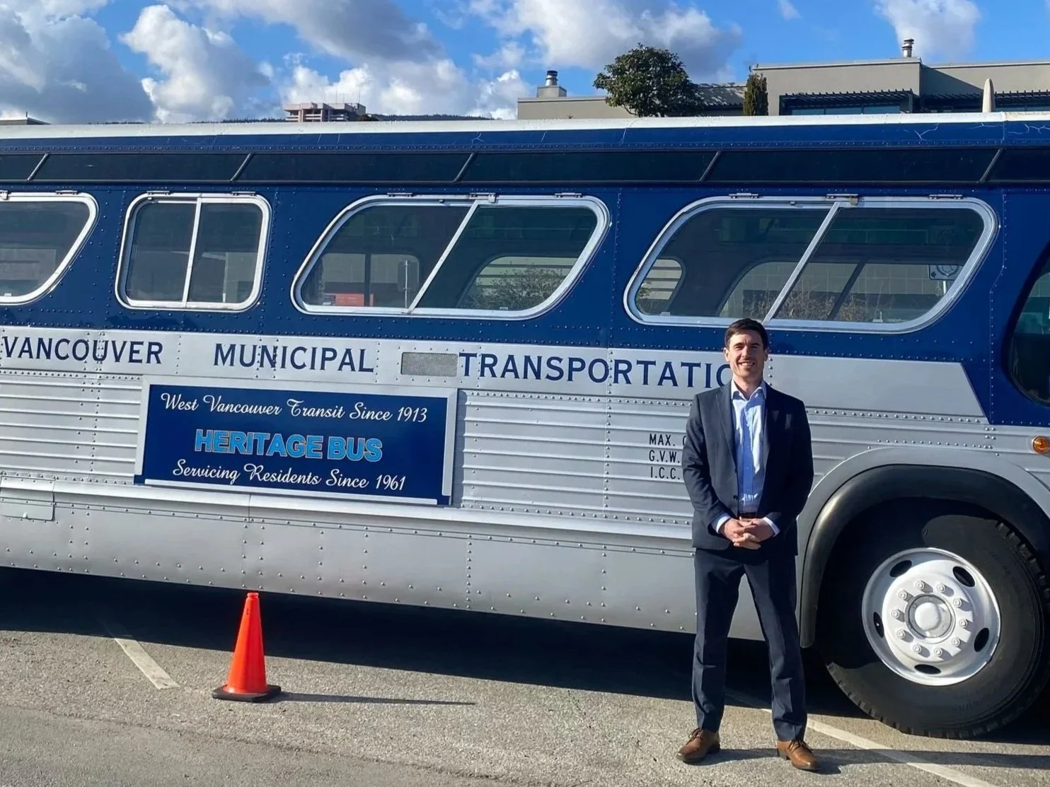 Man in a suit standing outside in front of a blue vintage bus with the sign 'Vancouver Municipal Transportation' and 'Heritage Bus' on the side, parked on a paved lot with a construction cone nearby and a blue sky with clouds overhead.