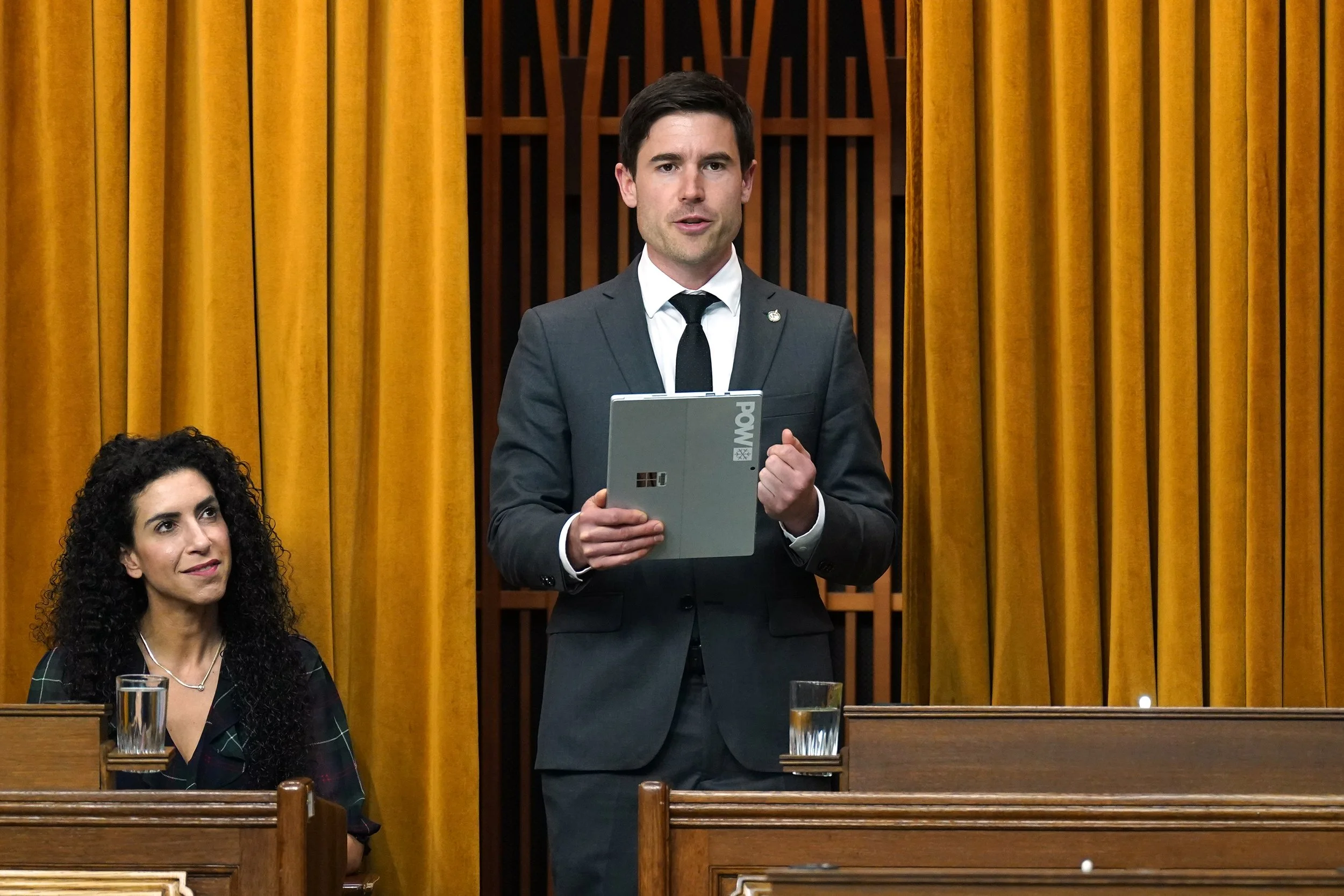 A man in a dark suit and tie stands holding a tablet in a formal setting with gold curtains behind him. A woman with curly dark hair sits nearby, also in formal attire.