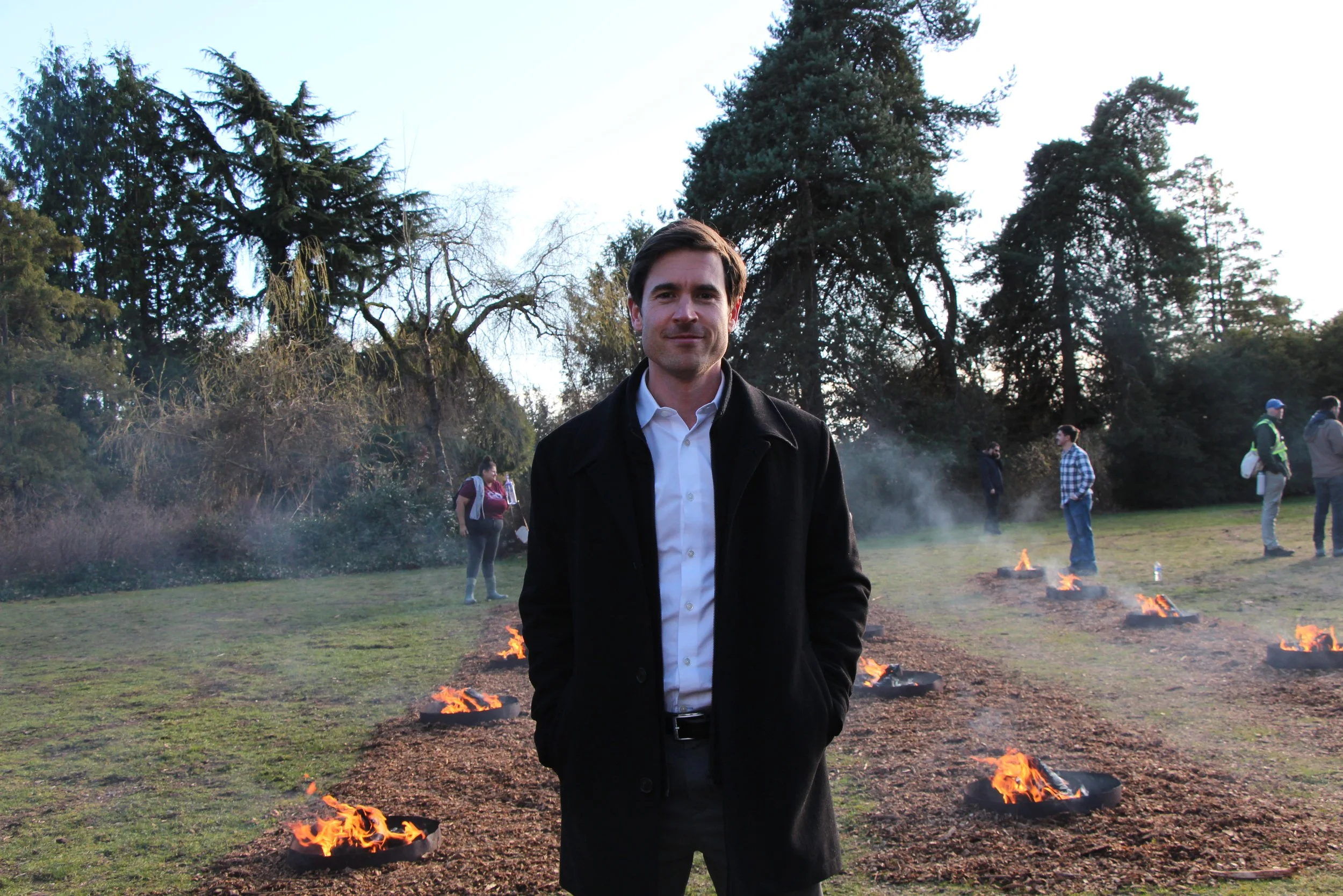 A man in a black jacket and white shirt standing outdoors in front of a line of small fires with trees in the background, and several people in the distance at a park.