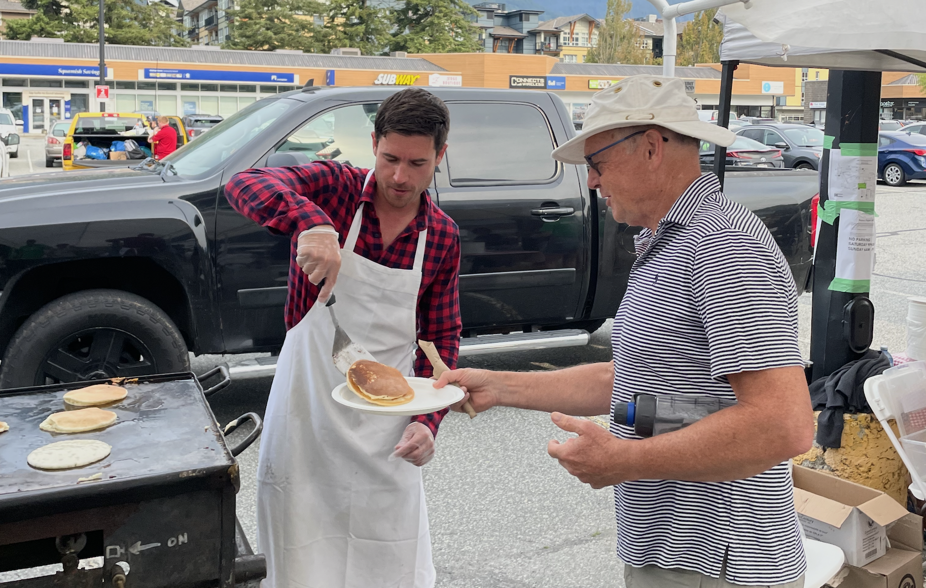A man in a striped polo shirt and white sun hat is receiving a plate of pancakes from another man wearing a red and black plaid shirt, white apron, and glove at an outdoor food stand. There are pancakes on a griddle in the foreground, and a parking lot with cars, a black truck, and a shopping plaza with various stores including Subway in the background.