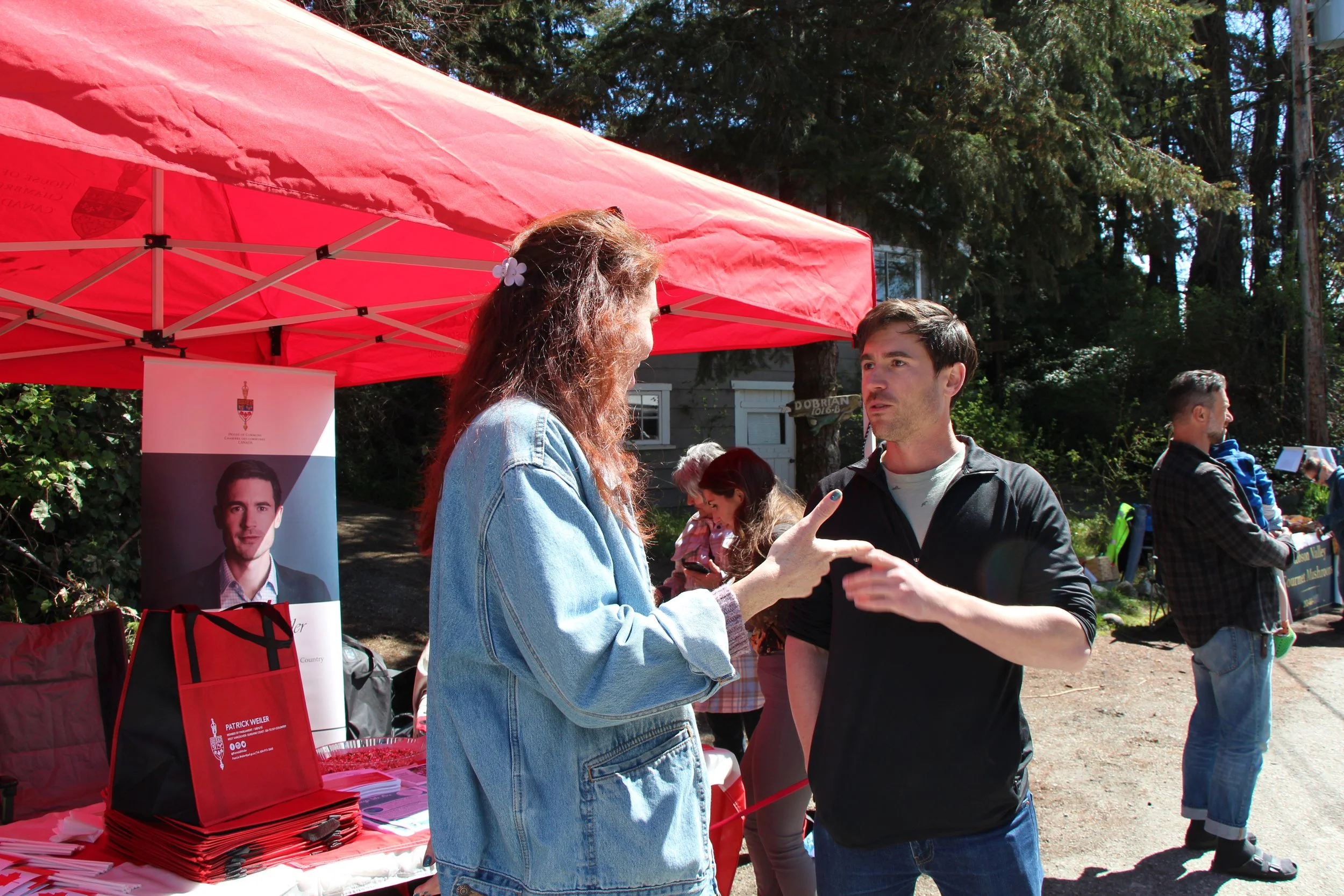 Two people are having a conversation at an outdoor table under a red canopy. There are flyers and brochures on the table, and a poster with a man's portrait and text is visible. Other people are in the background in a wooded area.