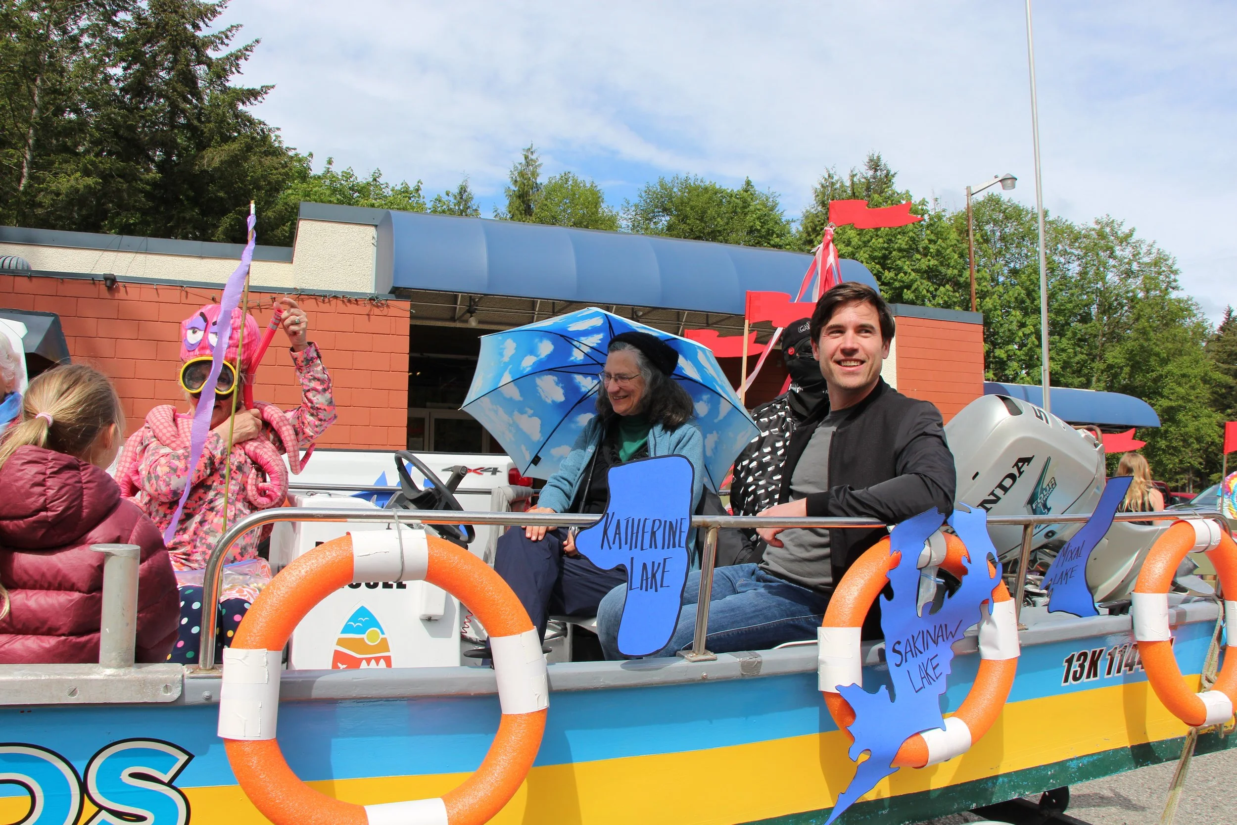 People on a boat decorated with colorful signs of lakes such as Katharine Lake, Sakinaw Lake, and Unaw Lake, participating in a parade or celebration outdoors on a partly cloudy day with green trees in the background.