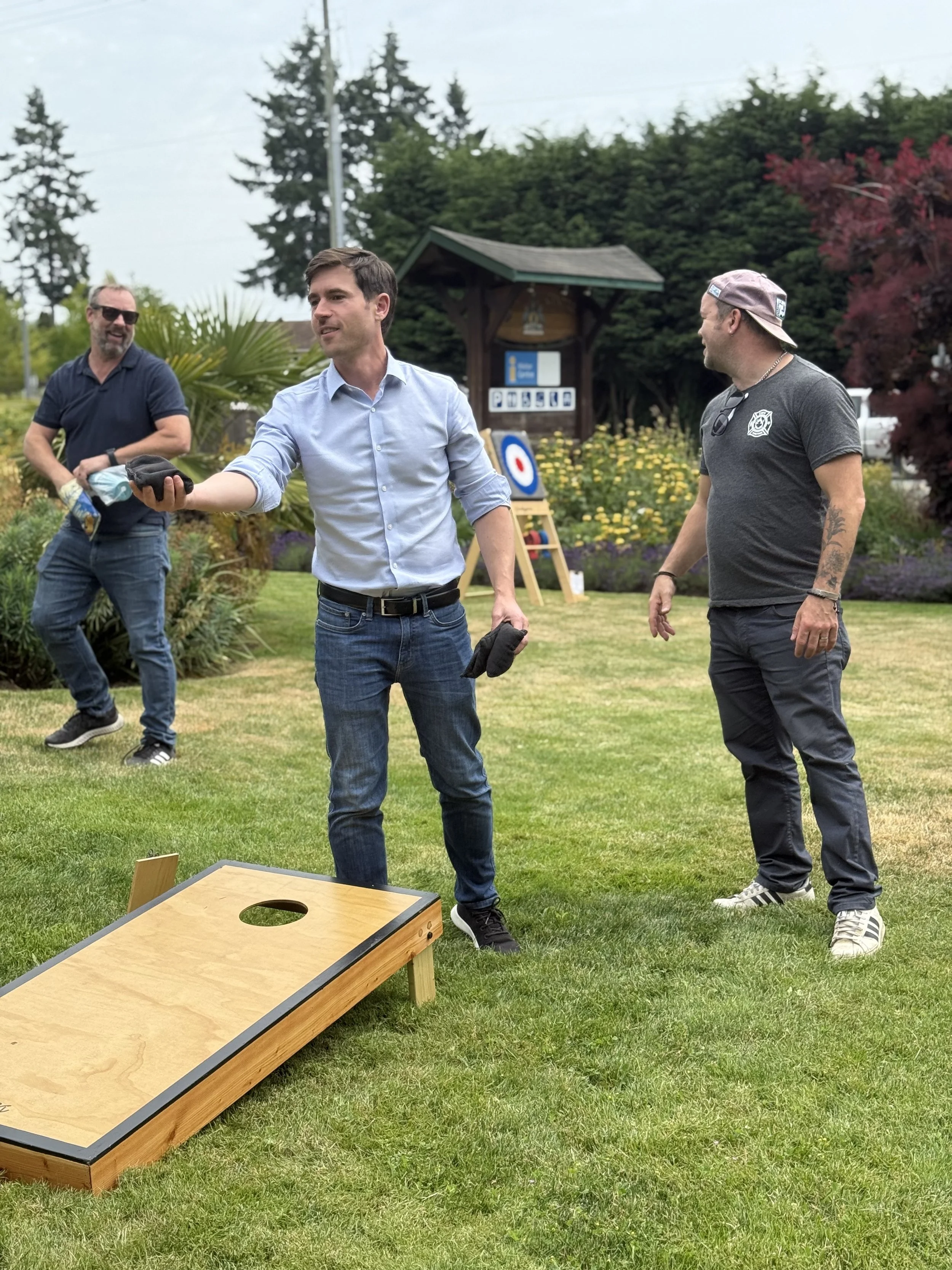 Three men playing cornhole outdoors in a grassy area, with trees, flowers, and a wooden sign in the background.