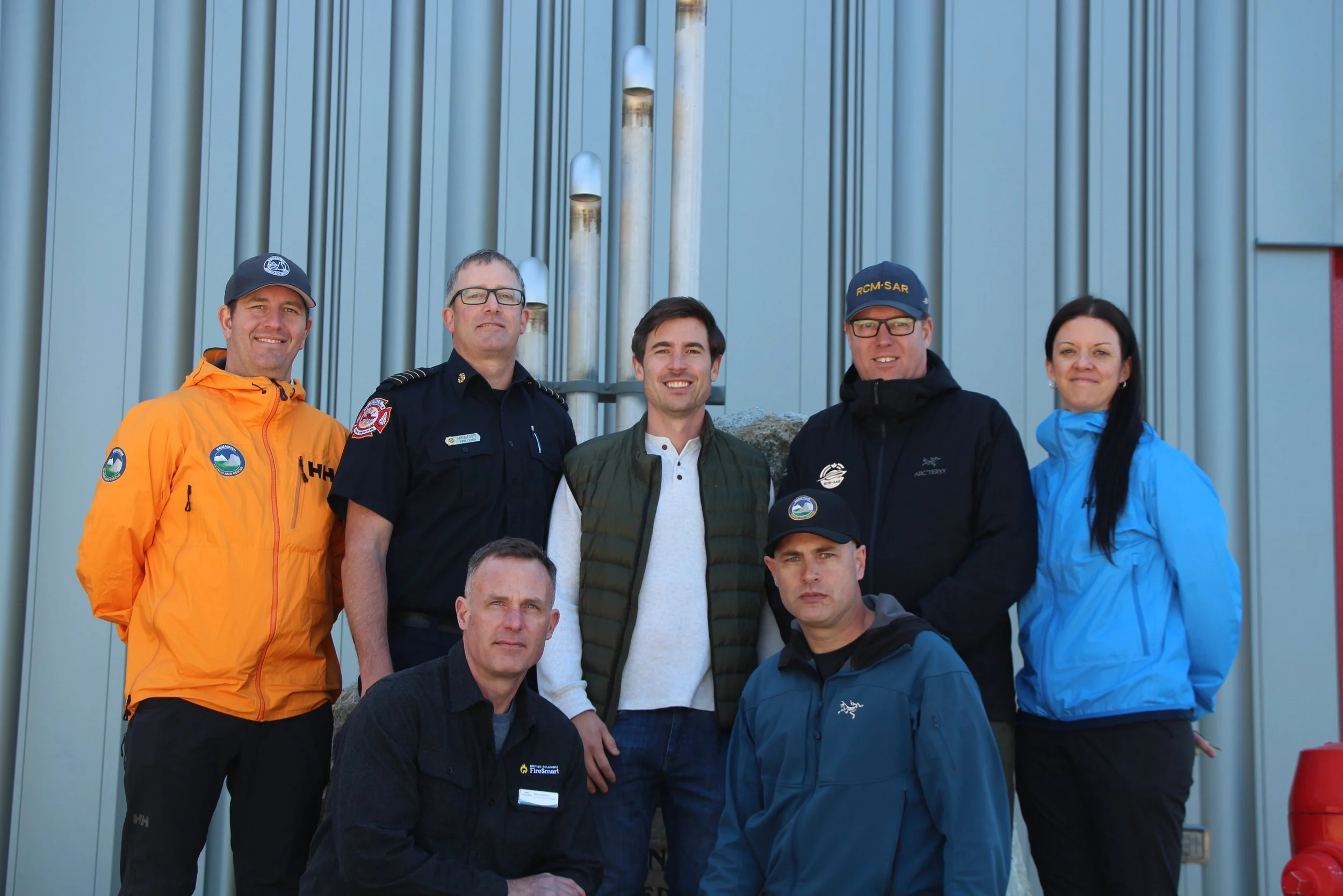 Group of six diverse people posing outdoors in front of a metal wall, some wearing uniforms related to emergency or rescue services.