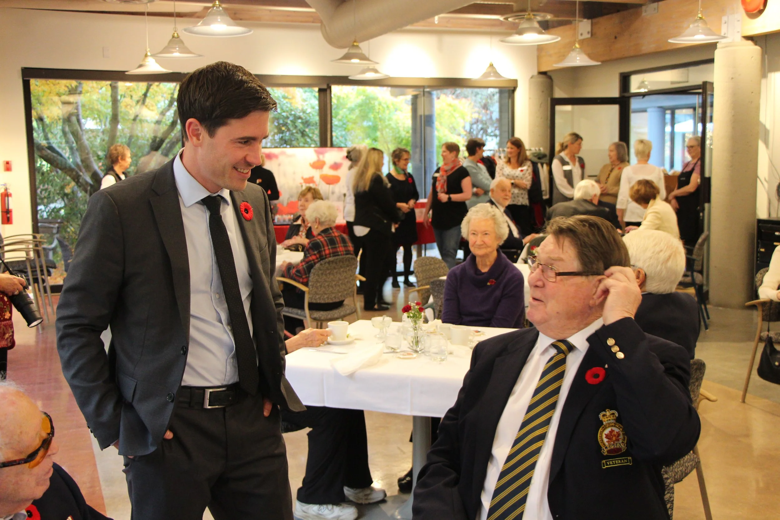A man in a gray suit and tie, smiling, talking to an older man in uniform with a poppy pin, in a communal dining setting with many seniors and staff, decorated with red poppies and large windows showing trees outside.