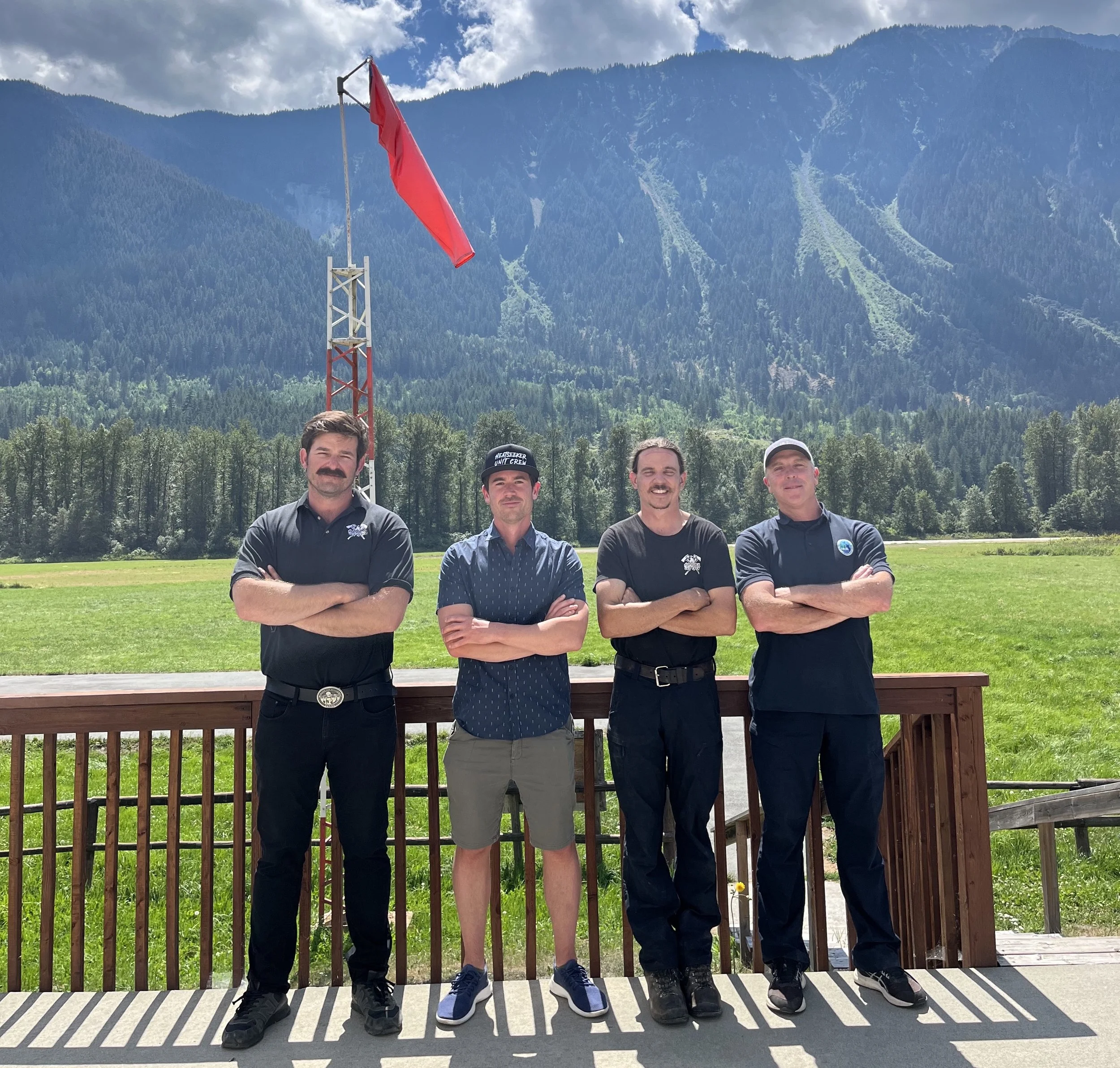 A group of five men standing outdoors on a wooden deck, with mountains and pine trees in the background. They are facing the camera with arms crossed, and a red windsock is visible behind them.