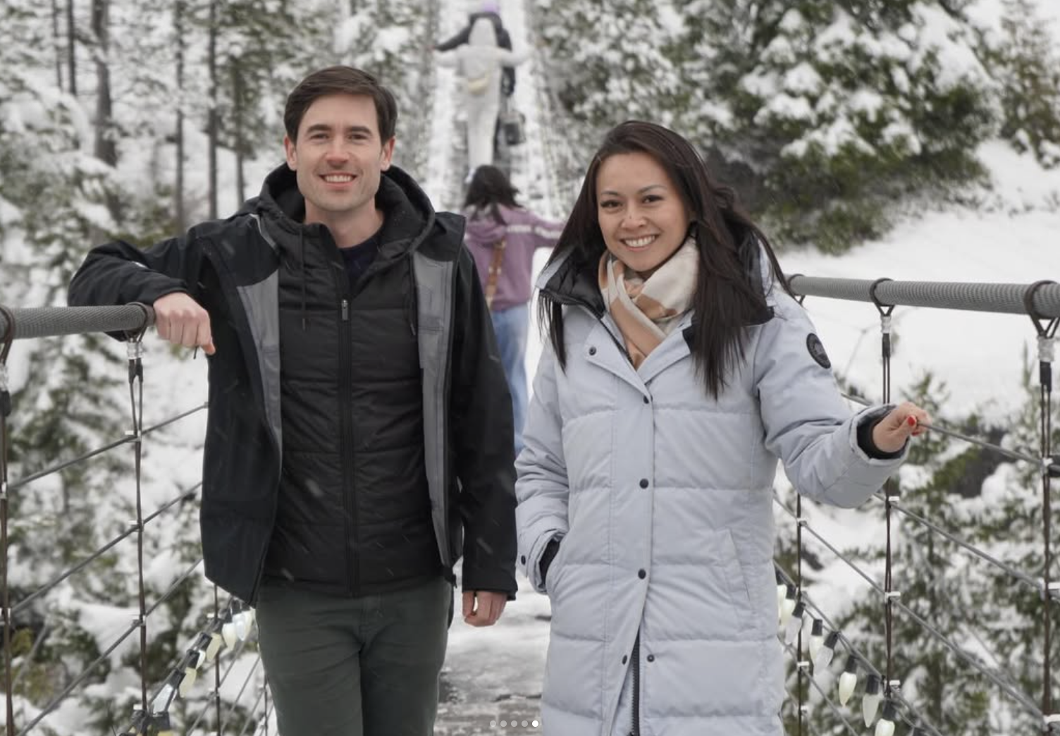 A man and a woman stand on a snowy suspension bridge, smiling at the camera, surrounded by snow-covered trees in a winter landscape.