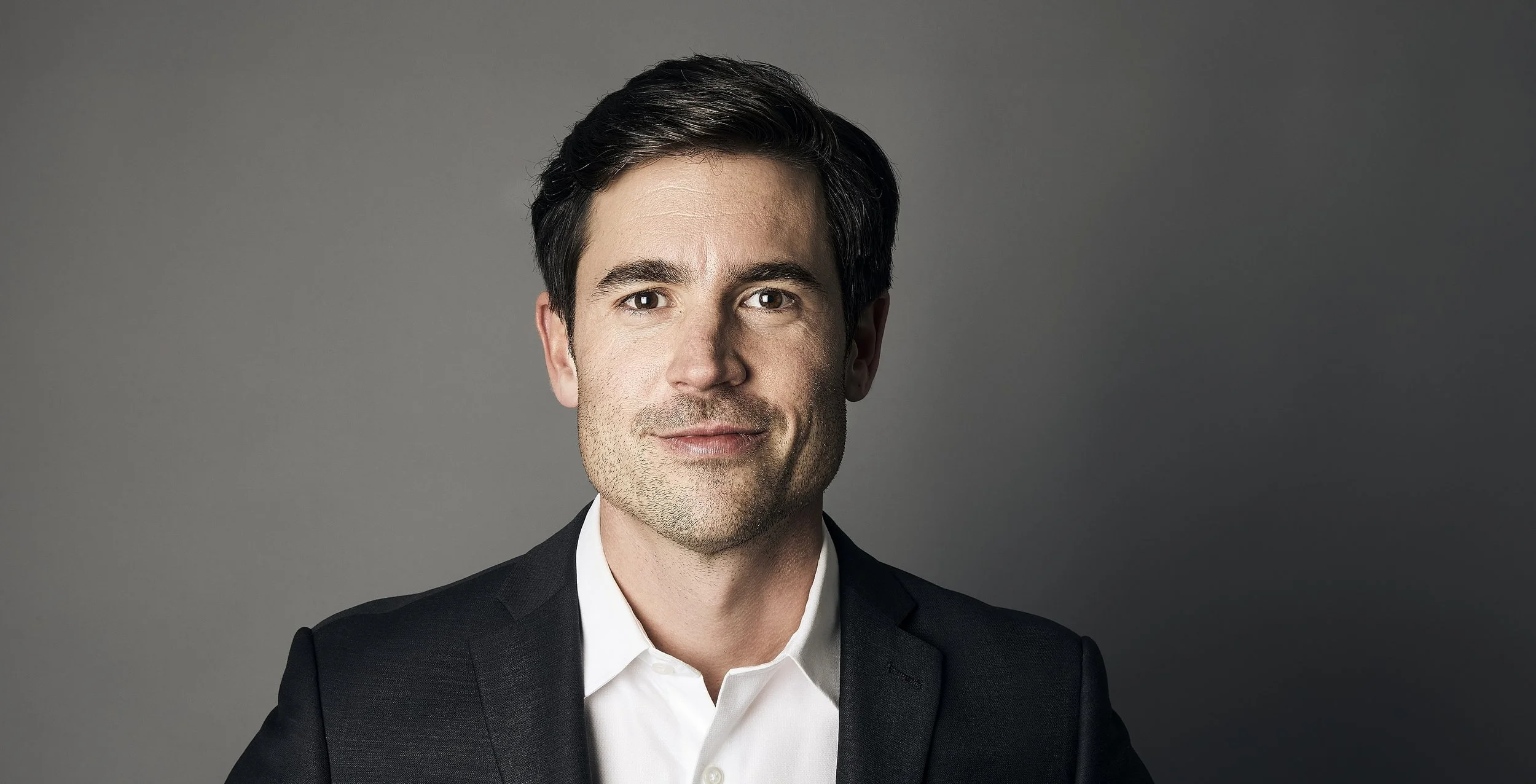 A professional man with dark hair and a slight smile, wearing a black suit and white shirt, against a gray background.