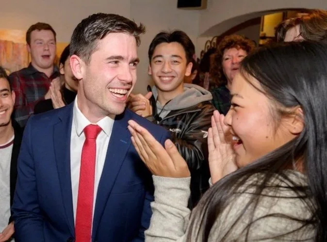 A man in a blue suit and red tie smiling as a woman facepalms while another woman touches his cheek, surrounded by a group of happy people in an indoor setting.