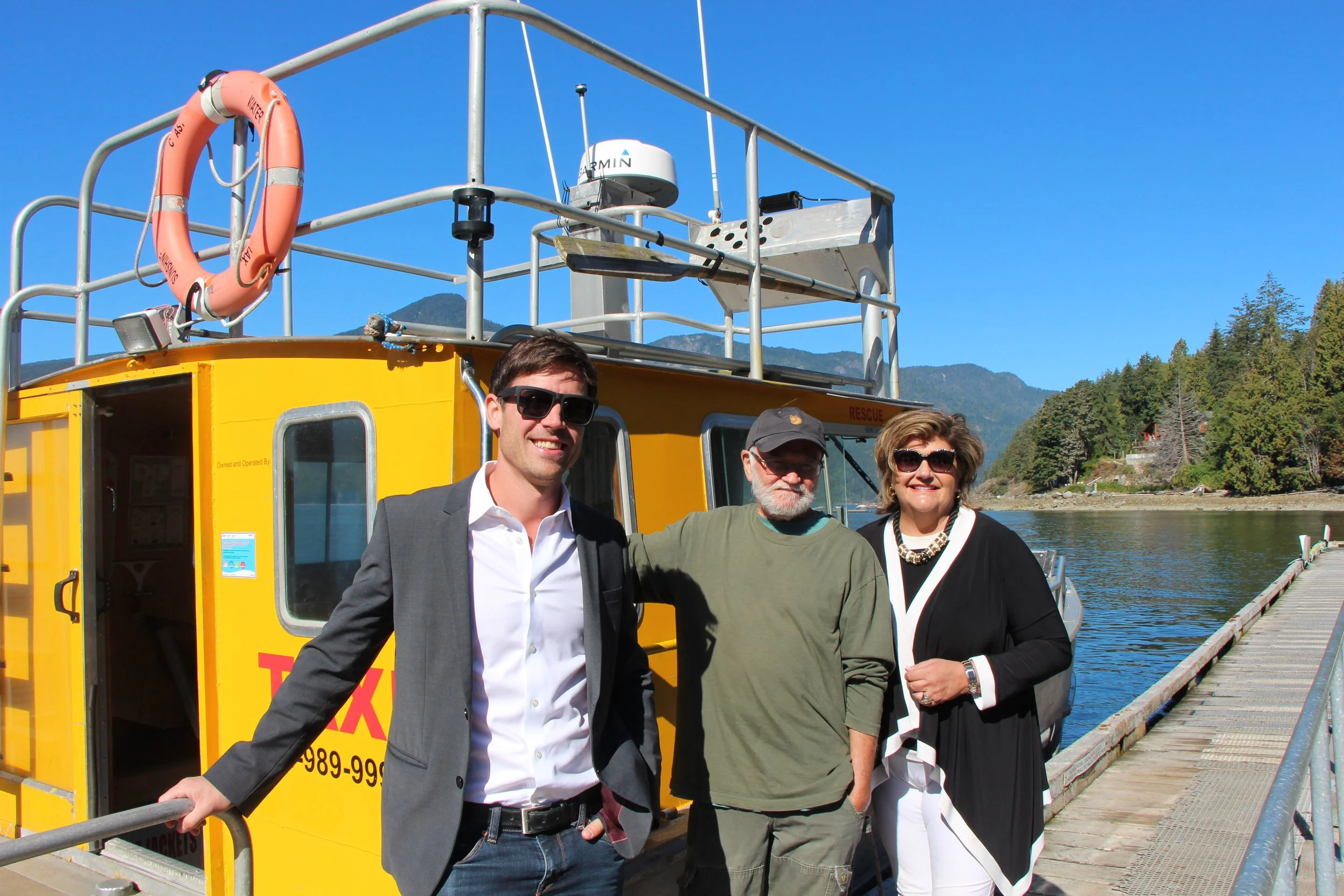 Three people standing on a dock beside a yellow boat with a railing and rescue equipment, with a mountain and trees in the background under a clear blue sky.