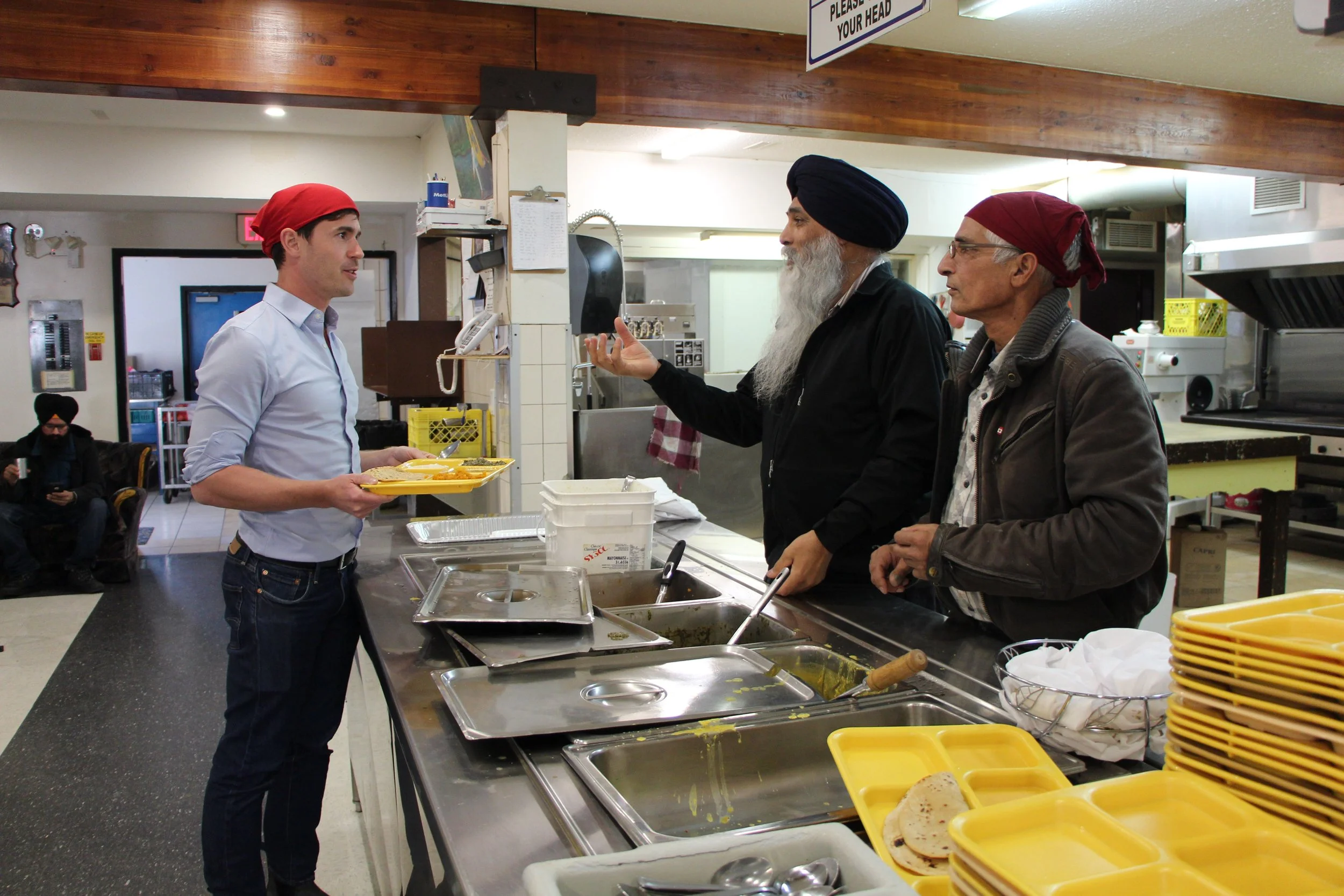 A young man wearing a red bandana receives food in a cafeteria from a man with a long beard and a black turban, while an older man with glasses and a red bandana looks on. The cafeteria has trays of food, yellow trays, and kitchen equipment in the background.