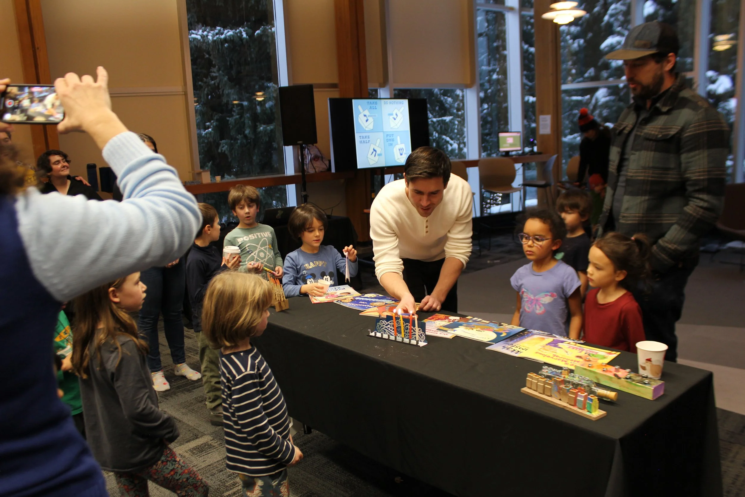 Children and adults gathered around a table with books and toys at a birthday celebration, with a man wrapping presents and a large screen displaying signs with instructions.