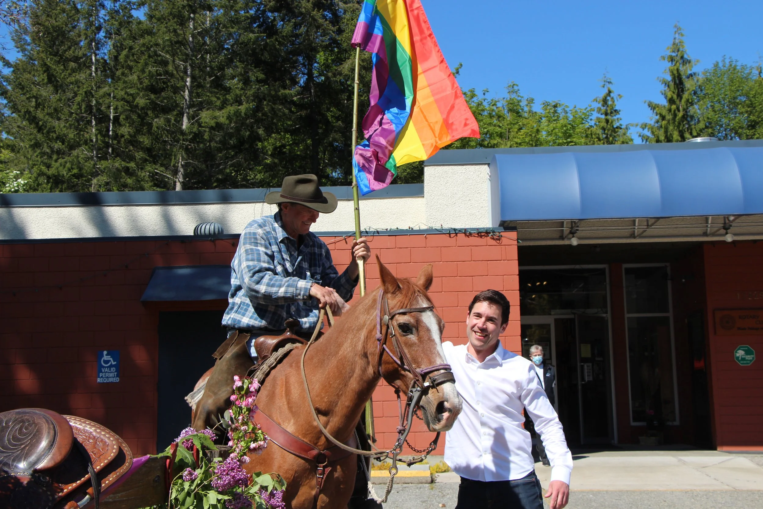 A man wearing a cowboy hat riding a horse decorated with purple flowers, with a man in a white shirt standing next to it. A rainbow flag is visible in the background, and a brick building with blue roof is behind them on a sunny day with green trees.