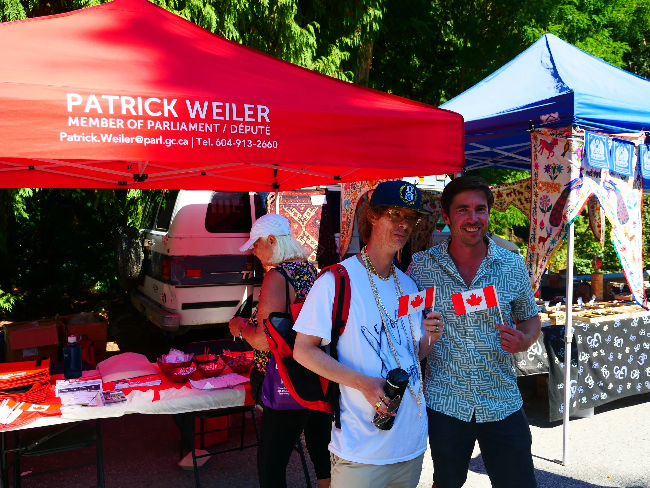 Two men smiling at an outdoor stall with red and blue tents, selling Canadian flags and patriotic items, with trees in the background.