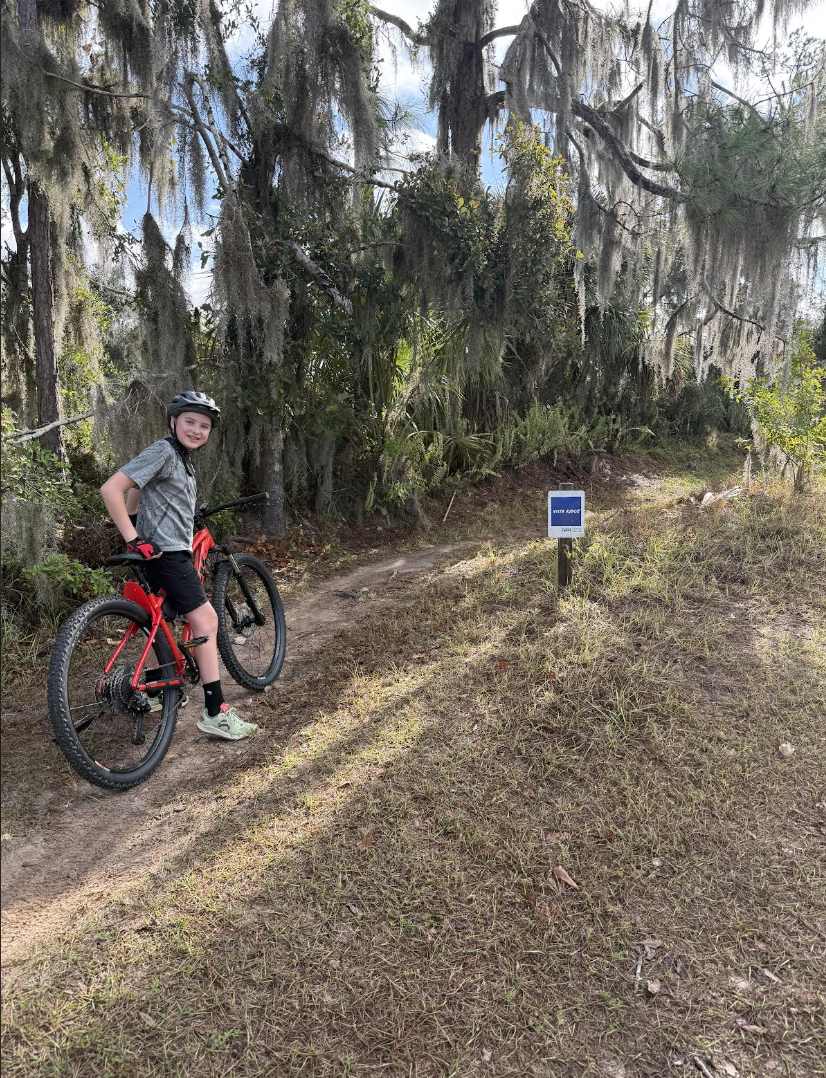 A young girl standing next to her red mountain bike on a dirt trail in a wooded area, wearing a helmet and summer clothes, with a smile on her face.