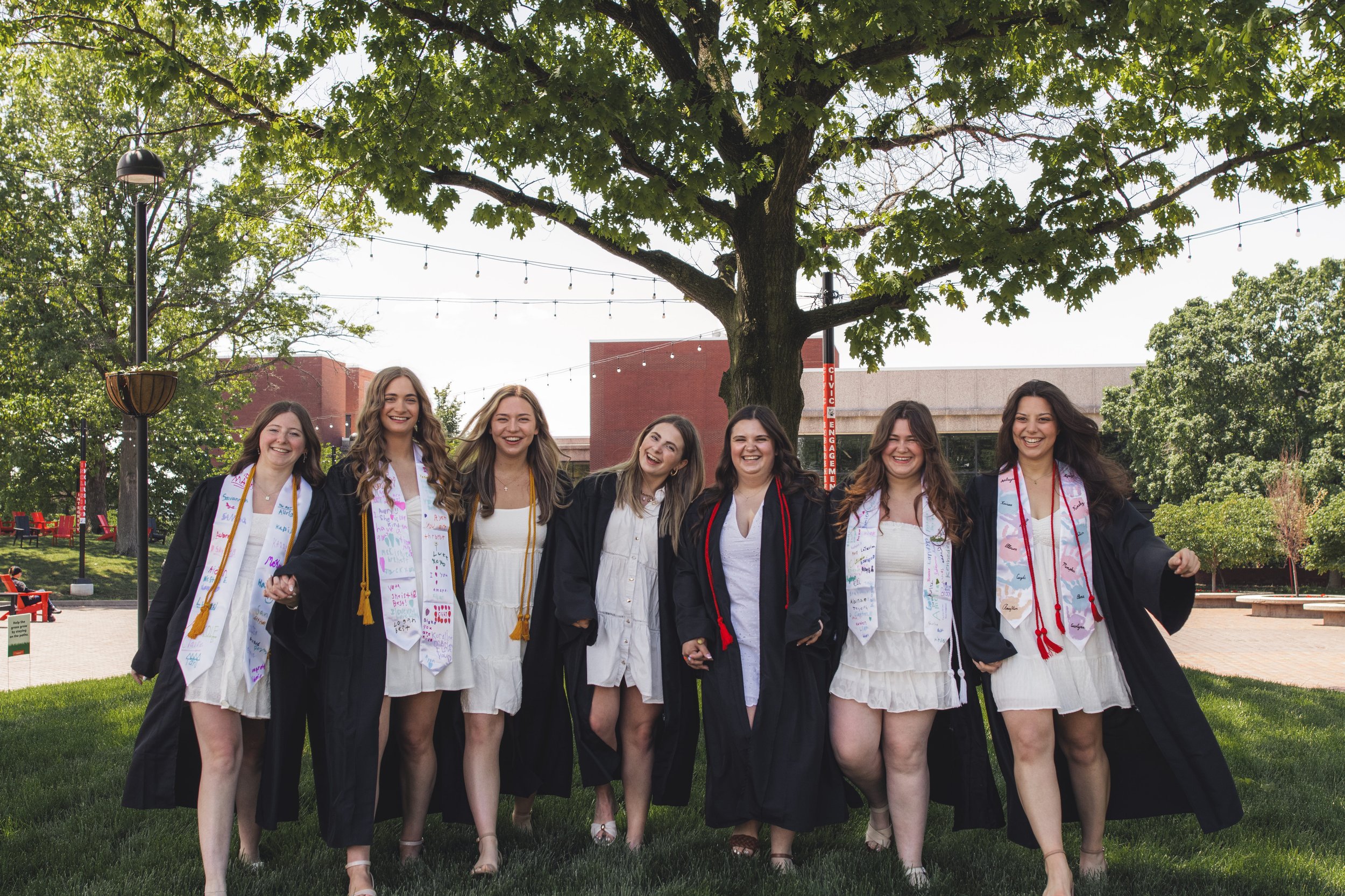 Group of young women in graduation gowns and sashes, smiling and holding hands outdoors under a large tree, celebrating a graduation.