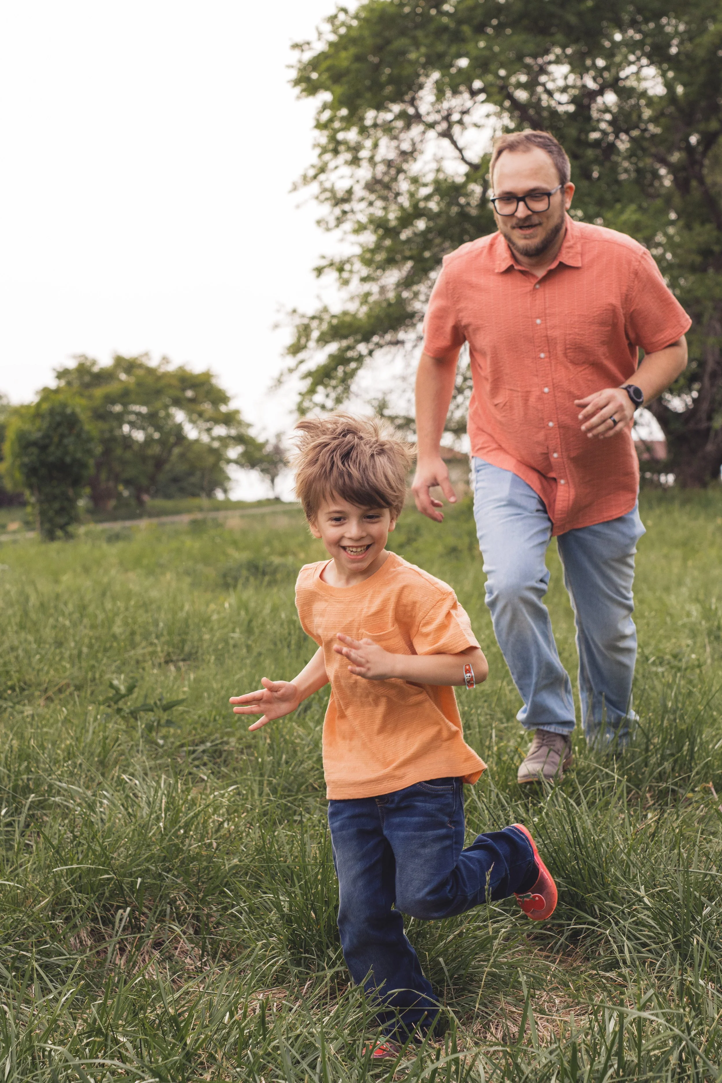 Father and son laughing and running together through a grassy field during a family photo session