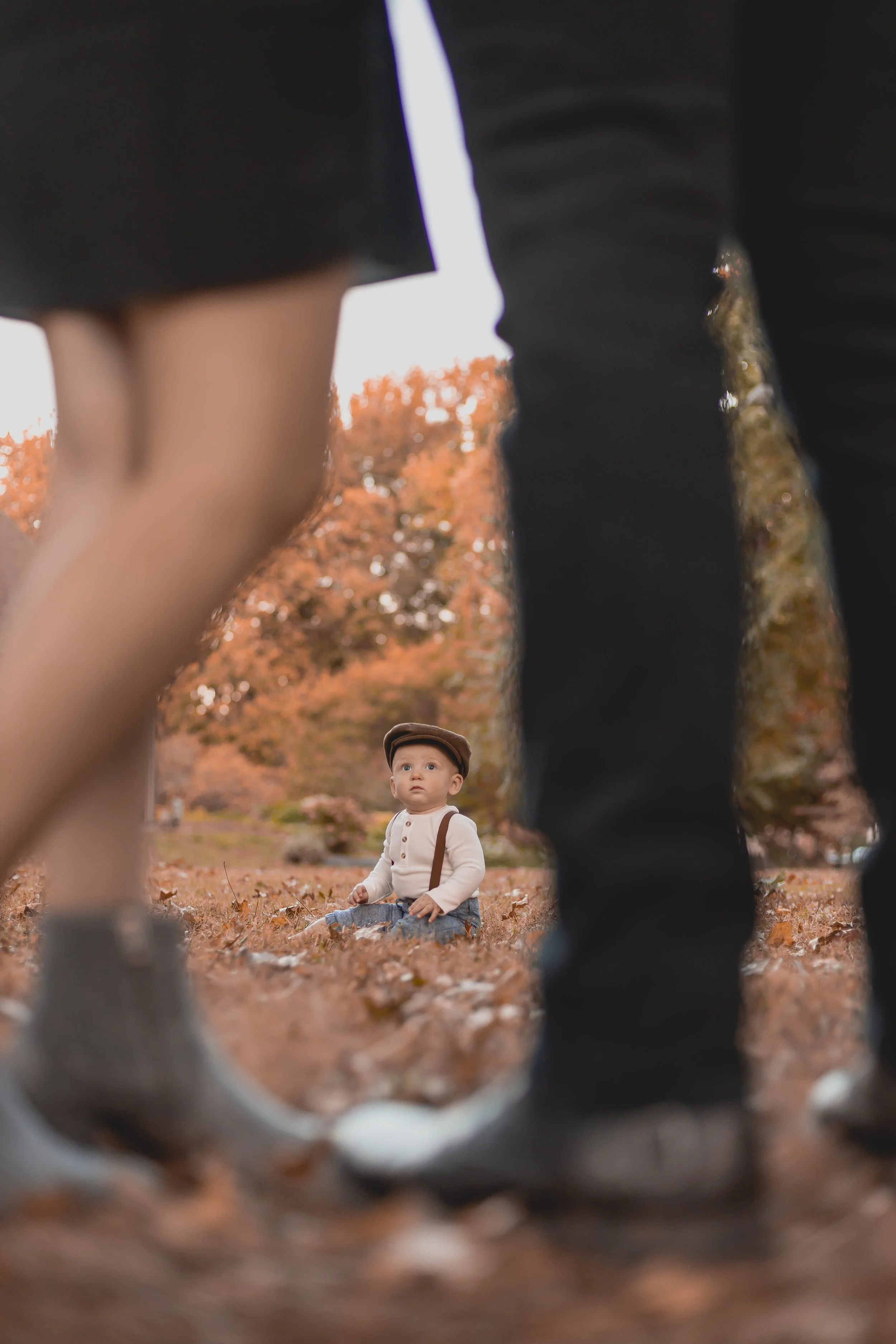 A young boy sitting on fallen autumn leaves in a park, viewed through the legs of his parents, with adults standing nearby and autumn trees in the background.