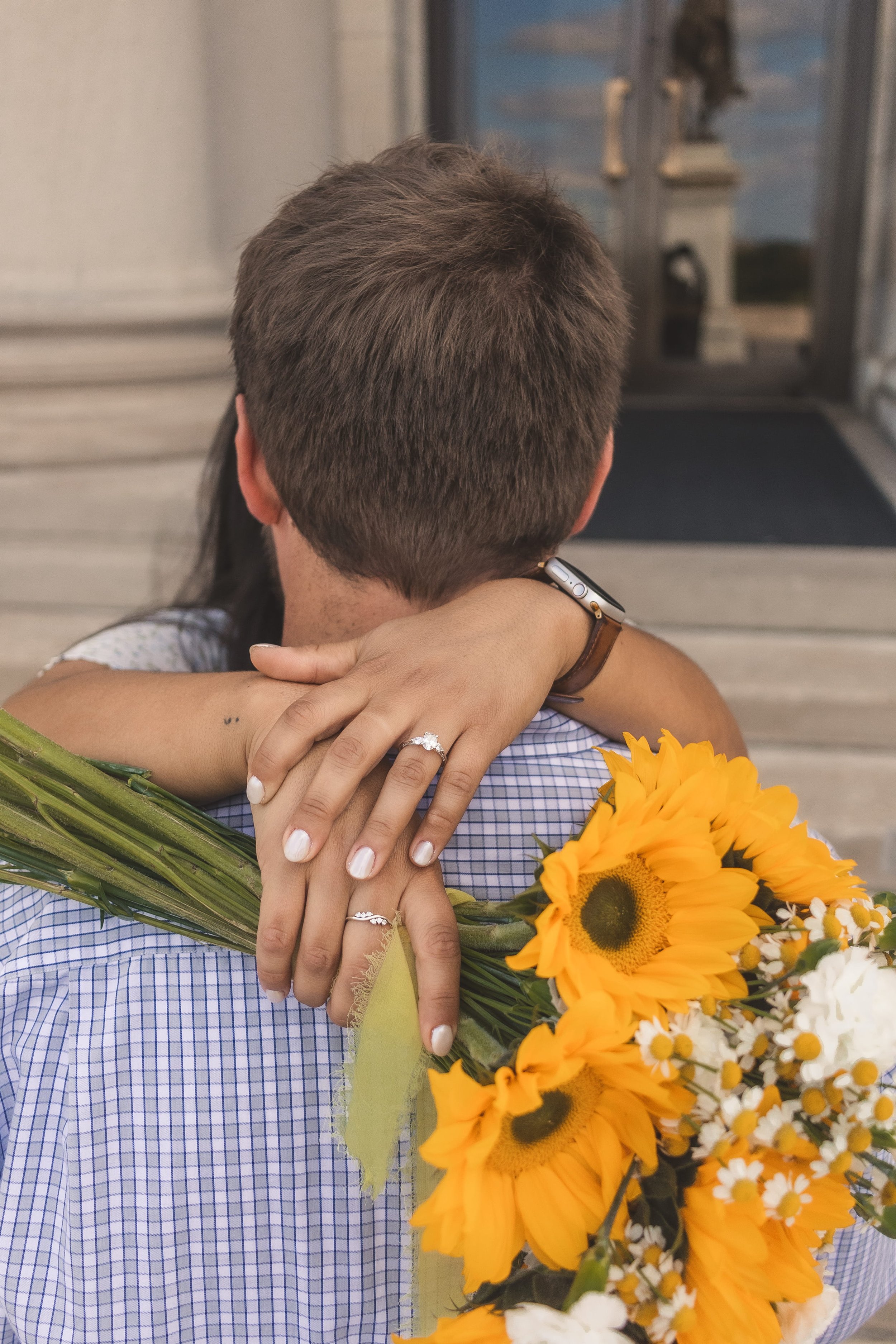 Engaged couple embracing with a sunflower bouquet, the woman's diamond engagement ring visible on her hand wrapped around her partner's shoulder