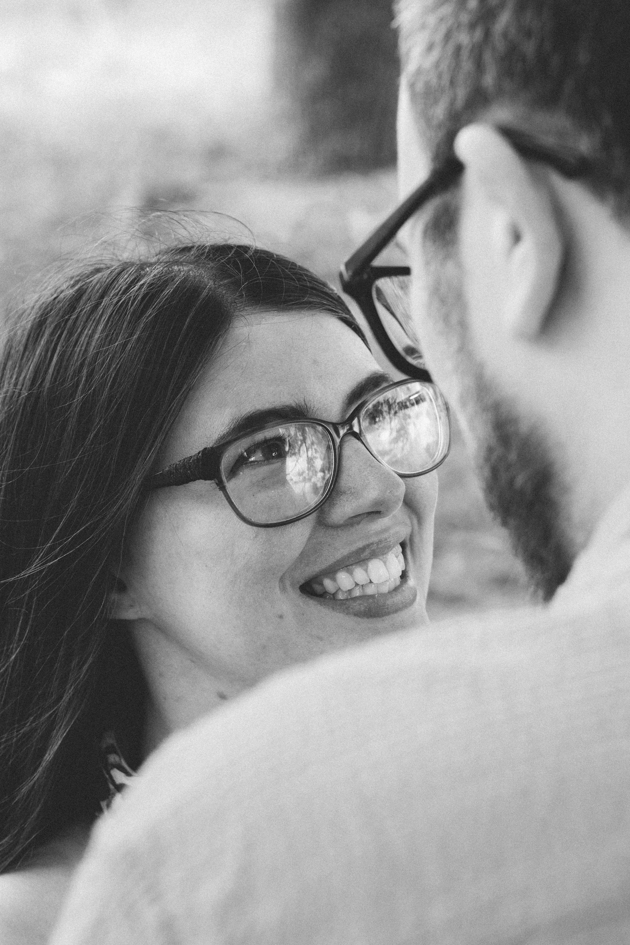 Close-up black and white photo of a woman with long dark hair and glasses, smiling and looking up, with a man close to her face in the background, also smiling and wearing glasses.