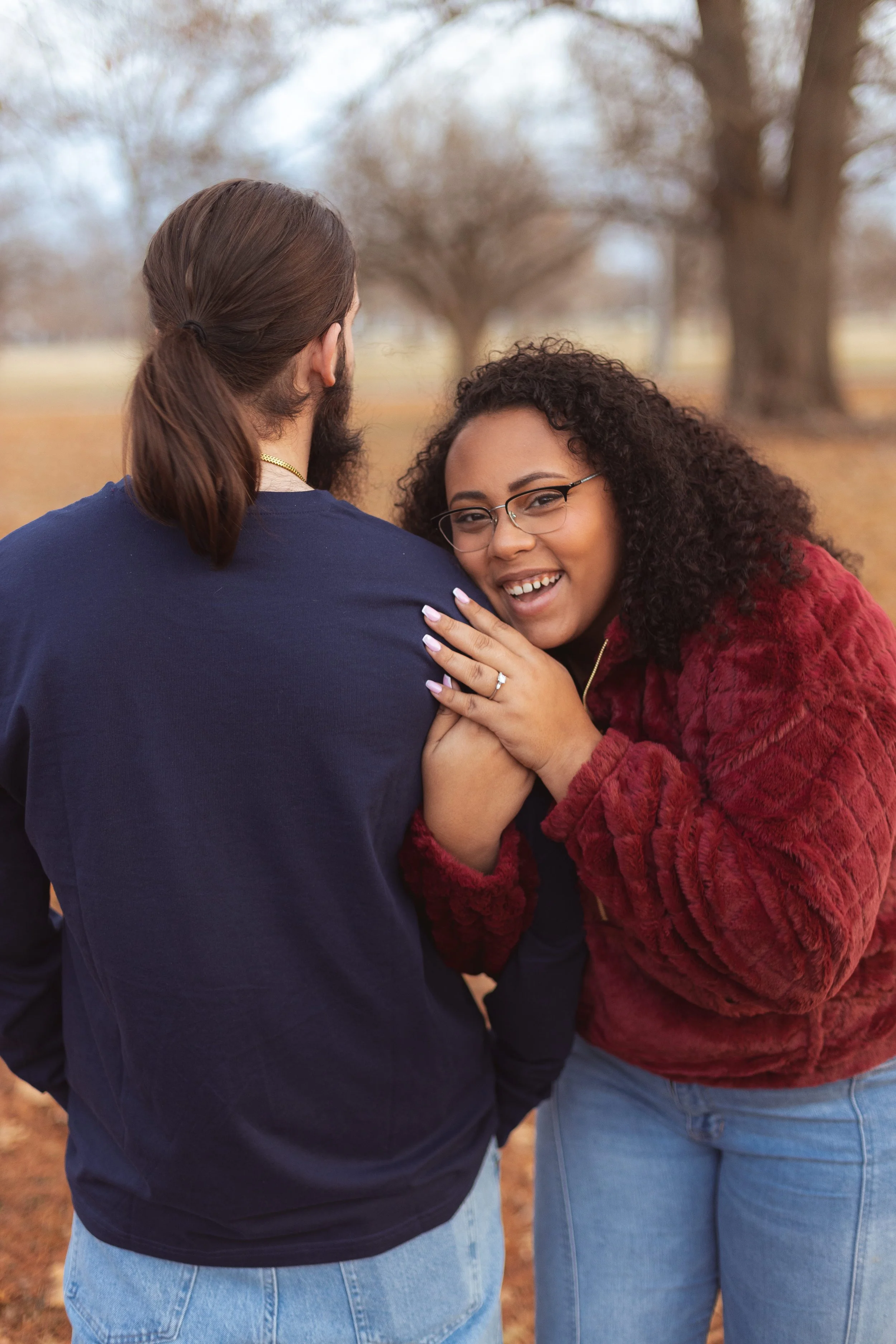A young couple embracing outdoors, one wearing a blue shirt and light blue jeans, the other in a red fuzzy jacket and light blue jeans, with trees in the background.