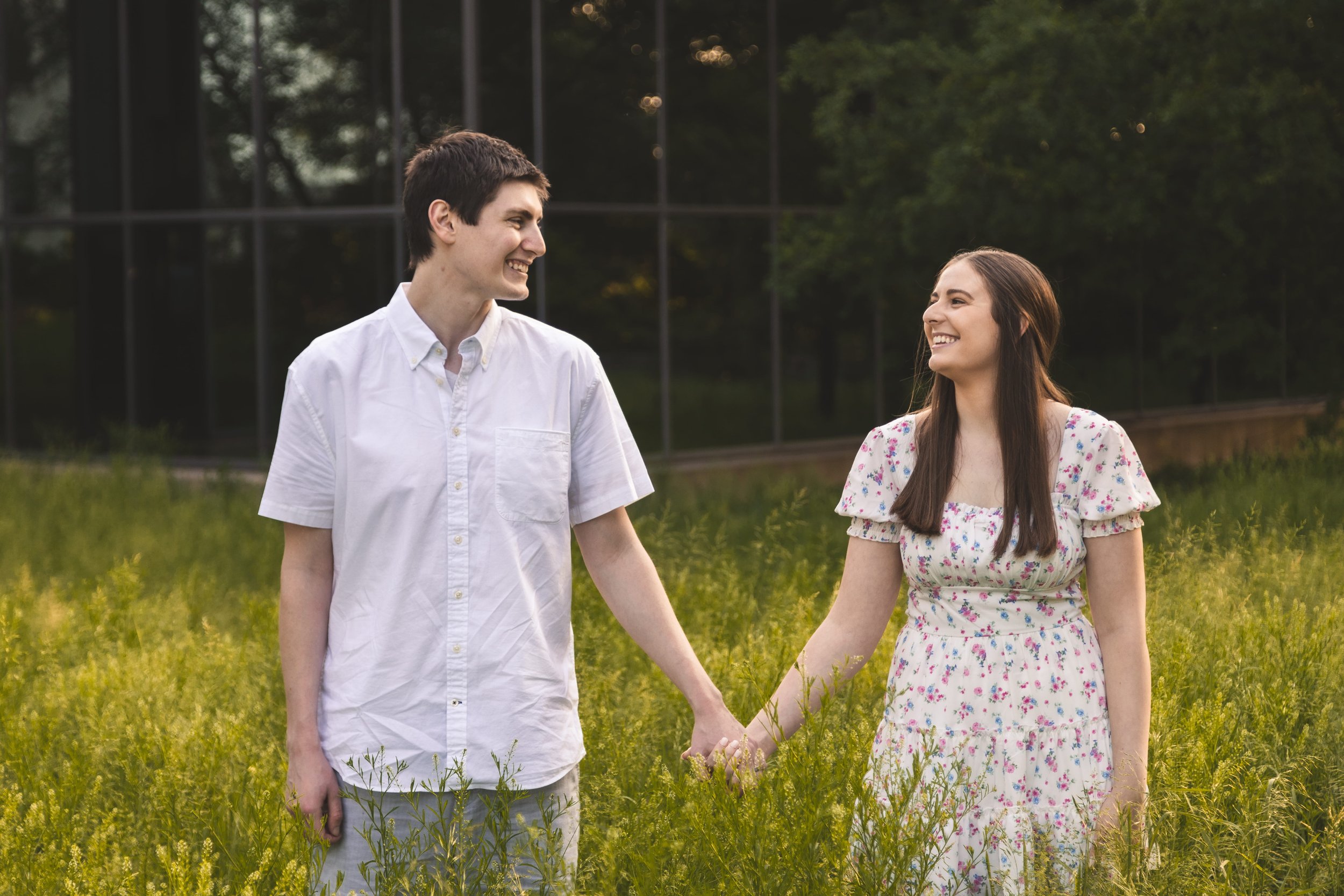 A young couple holding hands and smiling at each other while walking through a grassy field in the late afternoon or early evening.