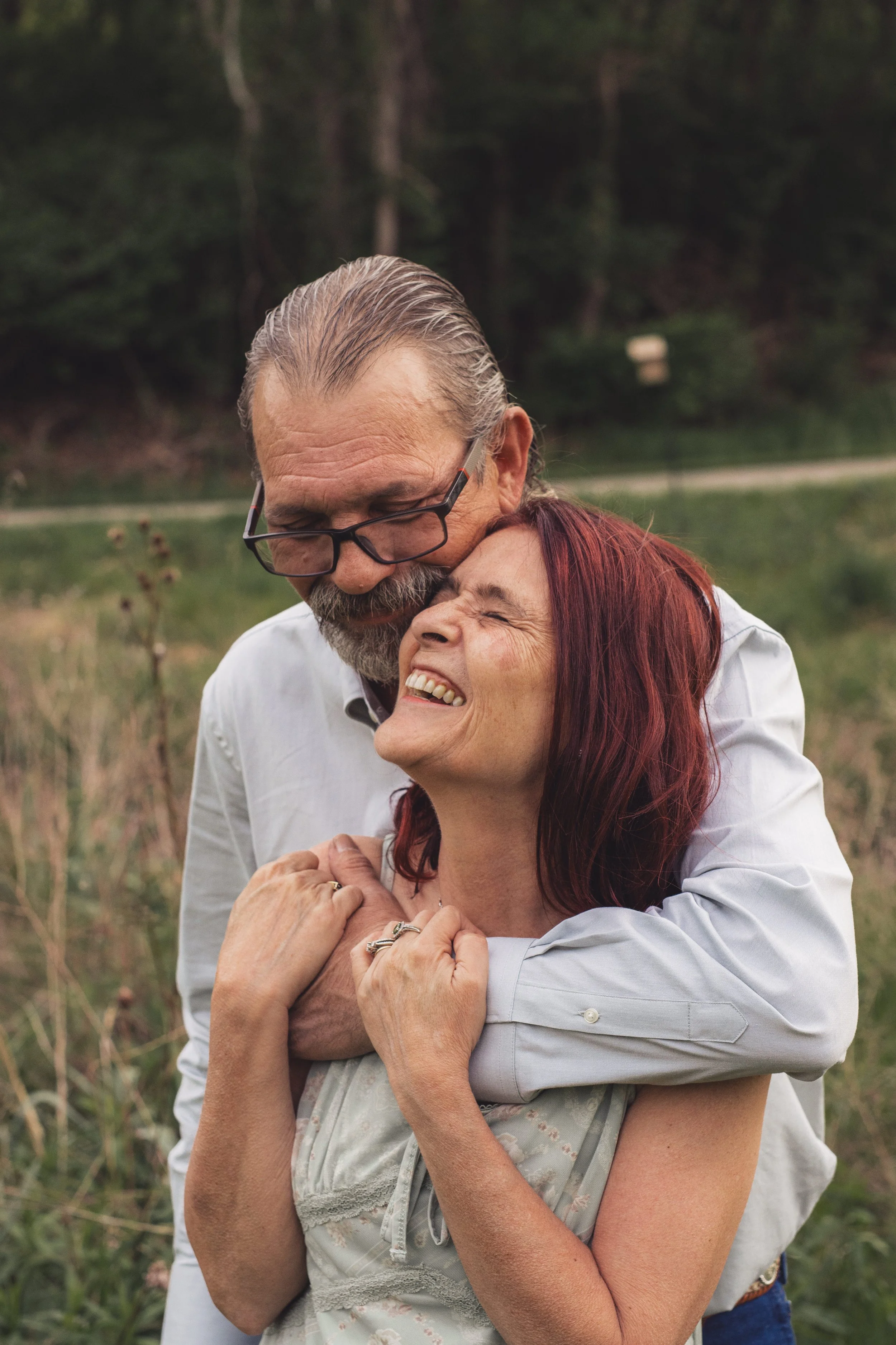 A man with glasses and a beard hugging a woman with red hair and closed eyes outdoors.
