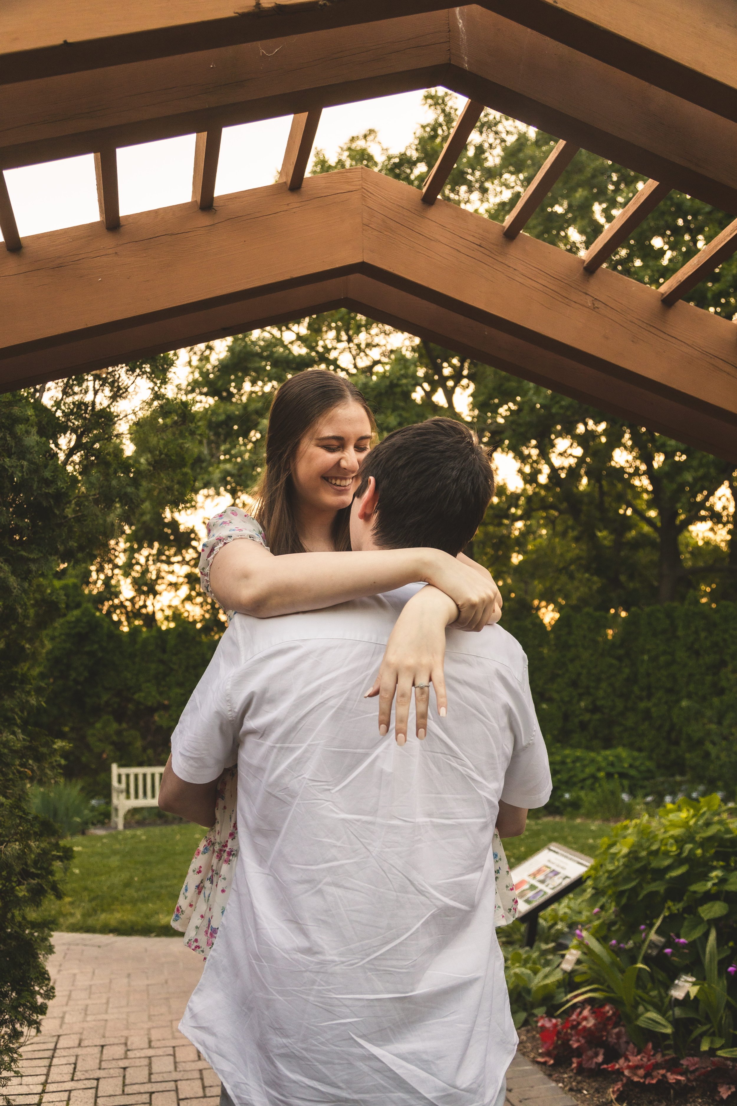 Couple embracing under a wooden pergola in a lush garden at golden hour, engagement photography