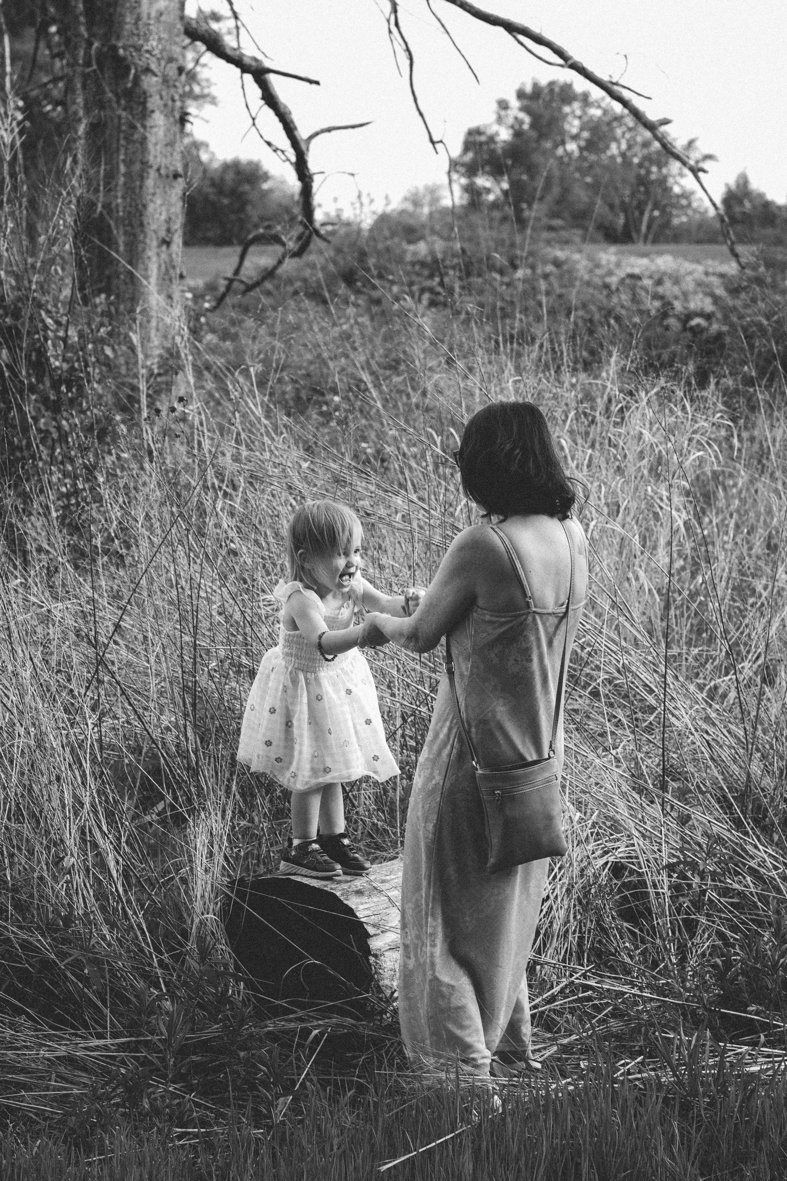A woman with shoulder-length dark hair and glasses, wearing a tank top and wide-legged pants, is holding hands with a young girl in a grassy field near a stream with trees and bushes in the background.
