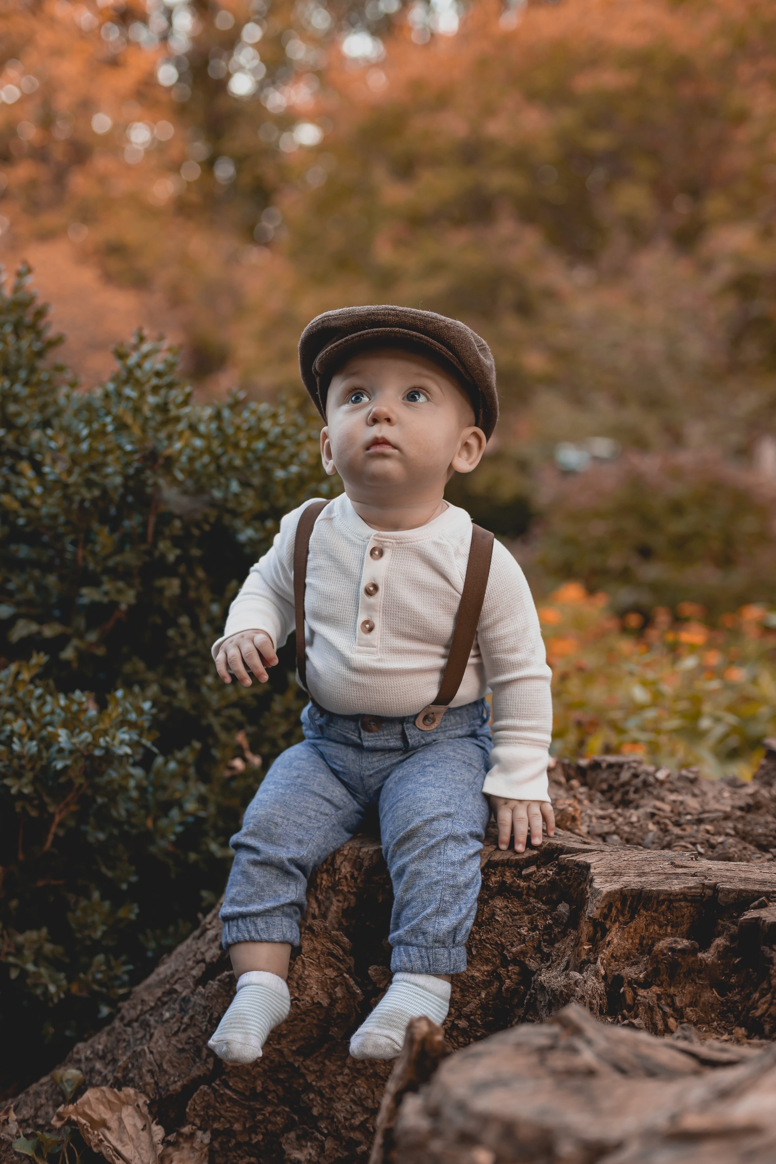 A young boy sitting on a tree trunk outdoors during autumn, wearing a brown cap, cream-colored shirt, blue jeans, and white socks, with colorful fall foliage in the background.