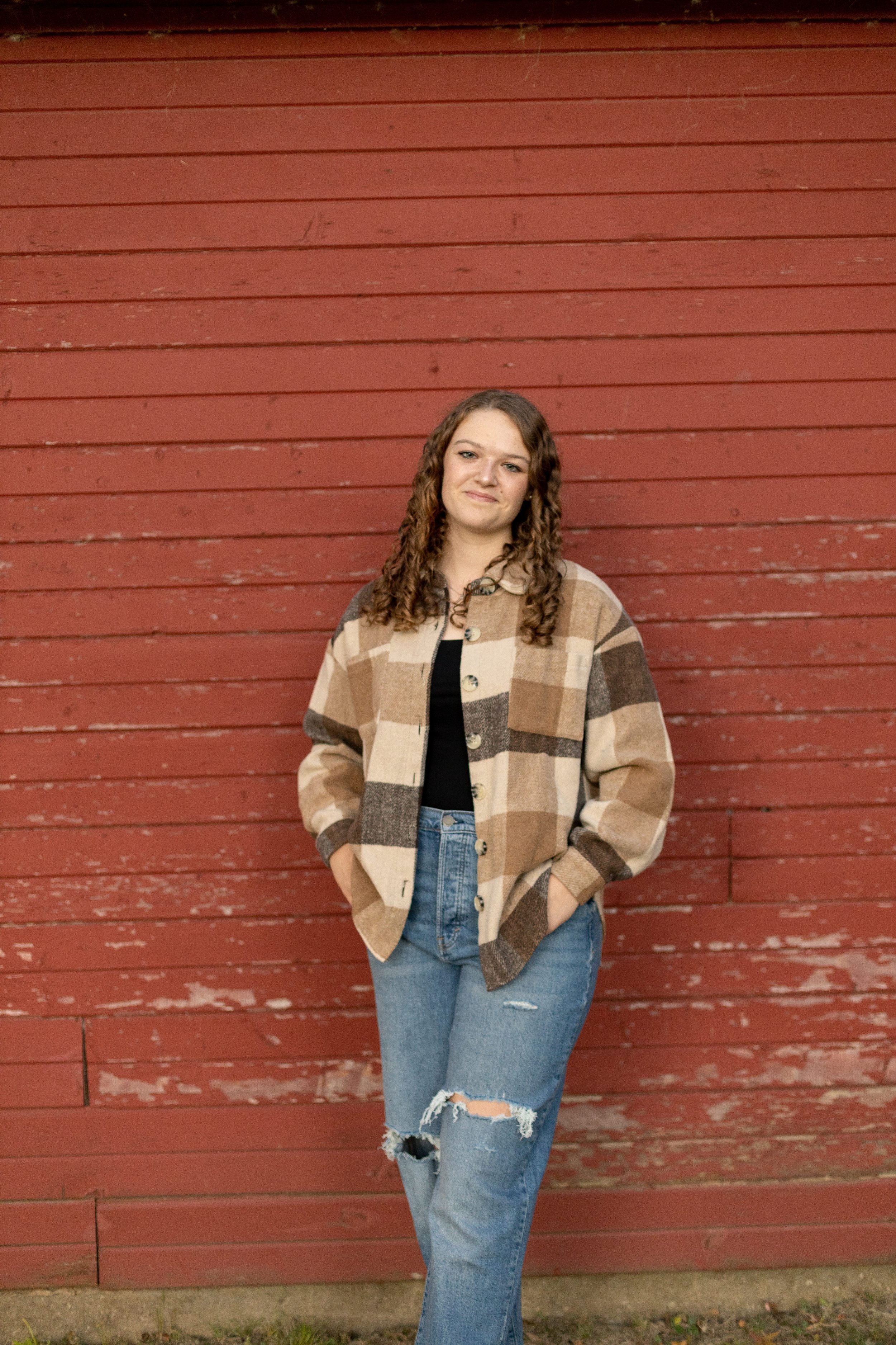 Young woman with curly hair wearing a beige and brown plaid jacket and ripped jeans standing in front of a red wooden wall.