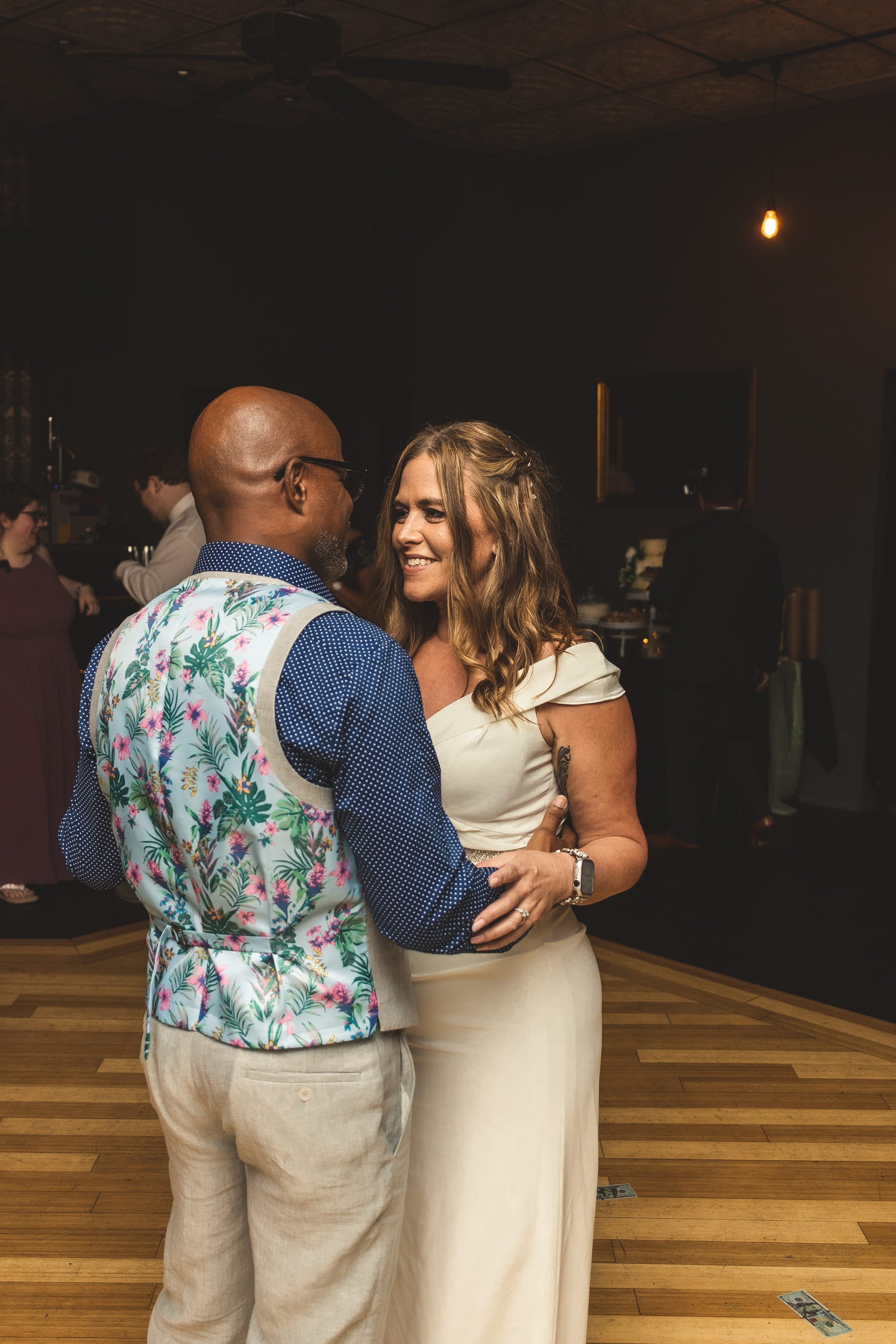 A couple sharing a dance at with other guests in the background. The man is bald, wearing glasses, a blue polka dot shirt, and a floral vest. The woman has long wavy hair, a light-colored dress. They are close, smiling, and looking at each other.