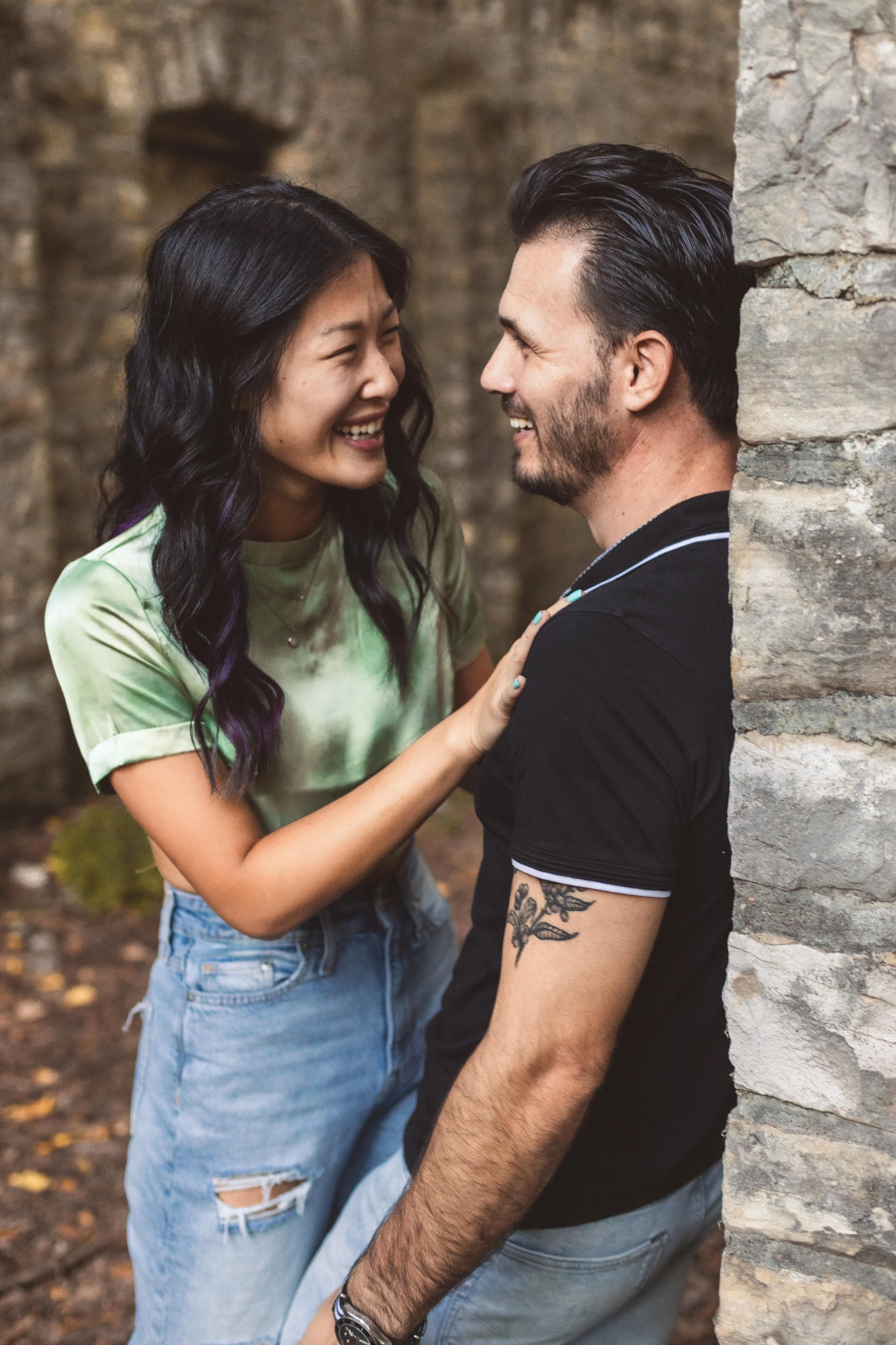 Couple laughing and sharing a joyful moment together during an outdoor engagement photo session near a stone wall