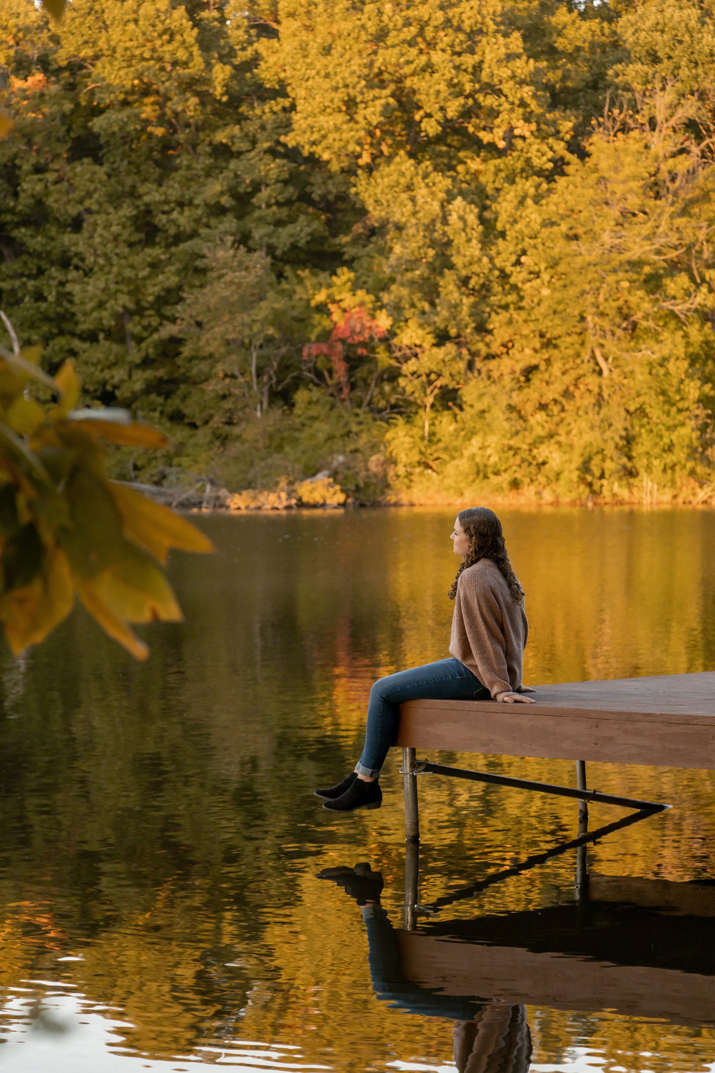 A woman sitting on a dock by a lake during autumn, with trees having yellow and orange leaves in the background.