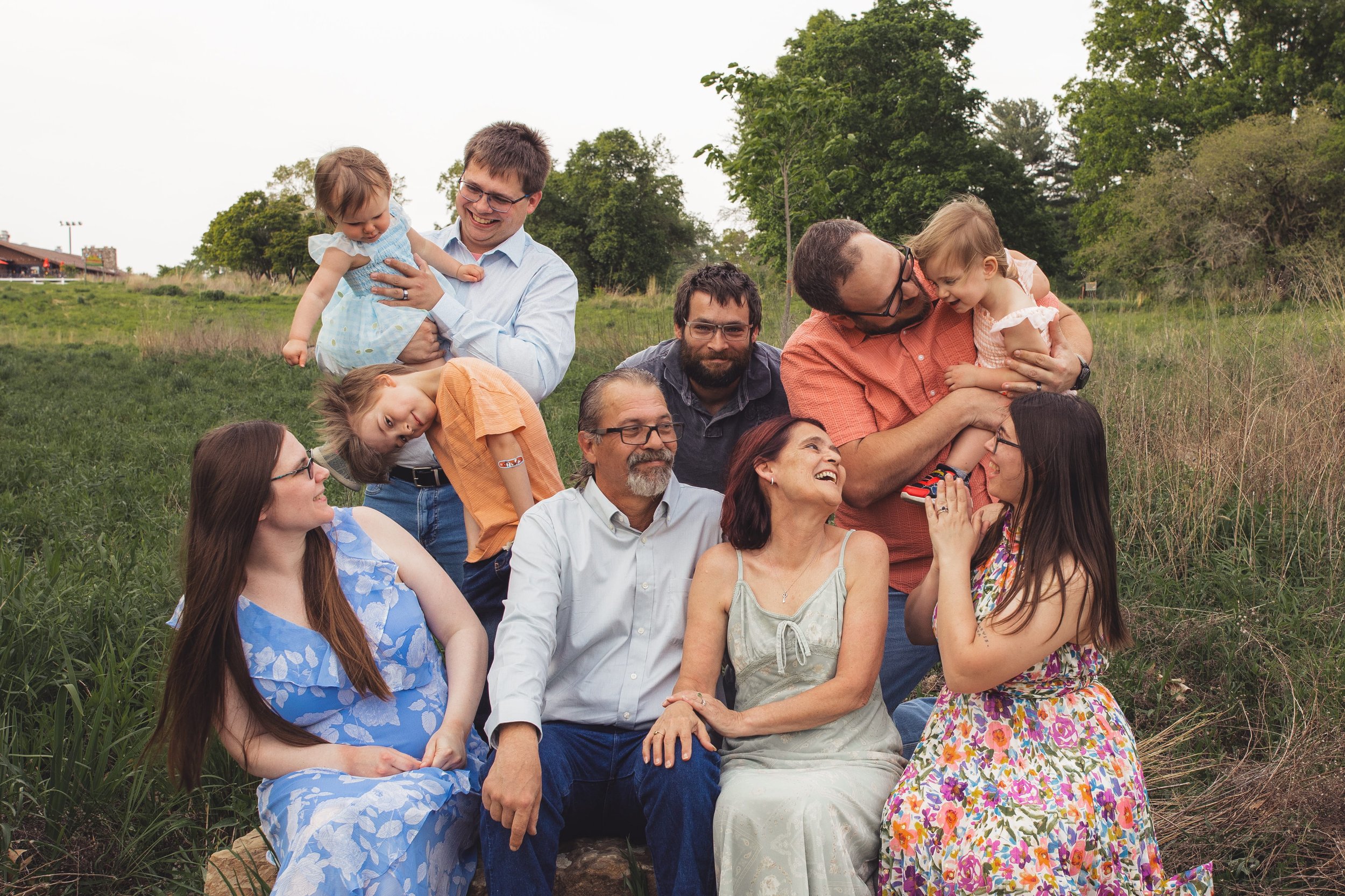 A group of adults and children outdoors in a grassy area, some sitting and some standing, smiling and hugging each other.